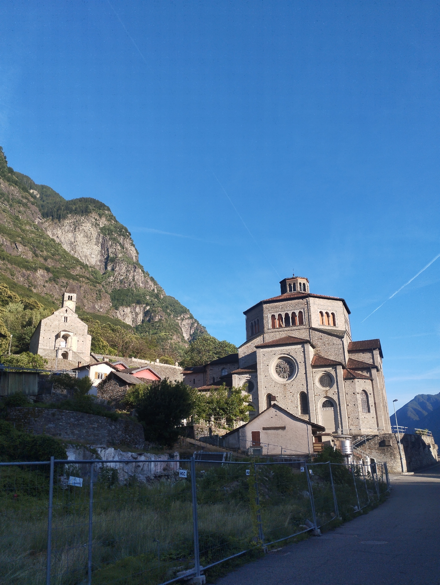 The image depicts a grand stone church with a distinctive octagonal dome, situated on a hillside with a steep, rocky mountain rising behind it. The church features arched windows, a large circular stained-glass window, and a tower. Smaller buildings, some with red roofs, are clustered around the church. In the foreground, a chain-link fence encloses an area with overgrown vegetation and rubble, indicating potential construction or renovation. A paved road runs alongside the fenced area, leading away from the church. The sky is a clear, bright blue with faint contrails from aircraft. The overall scene is bathed in natural daylight, suggesting it was taken during the day.
