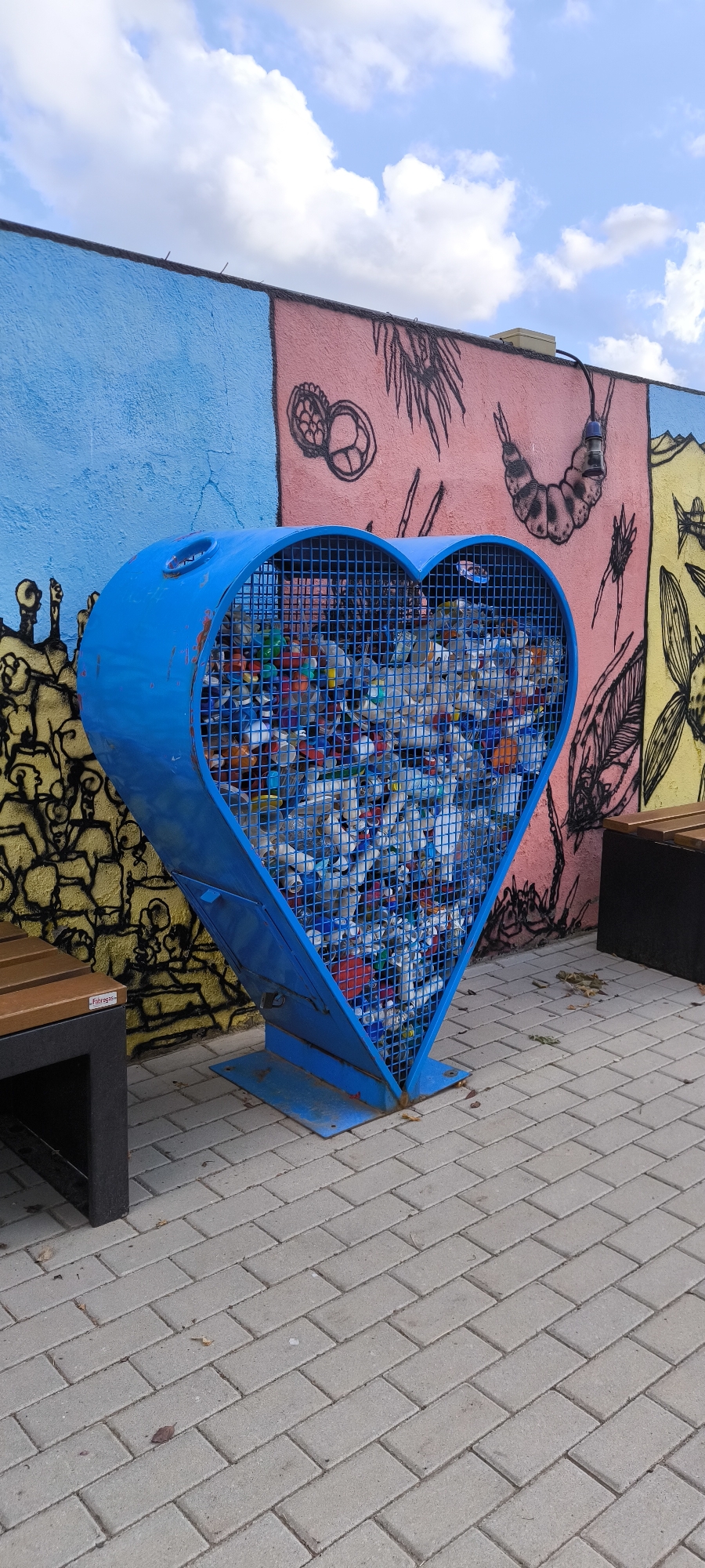 A blue, heart-shaped recycling bin, filled with plastic bottles, stands on a paved walkway in front of a brightly painted mural. The mural features various abstract and figurative drawings in pink, yellow, and blue. To the left and right of the bin are dark-colored benches with wooden slats. The ground is covered with rectangular gray pavers. The sky above is blue with scattered white clouds. The location is identified as Fanzara, Spain. There are no people visible in the image.