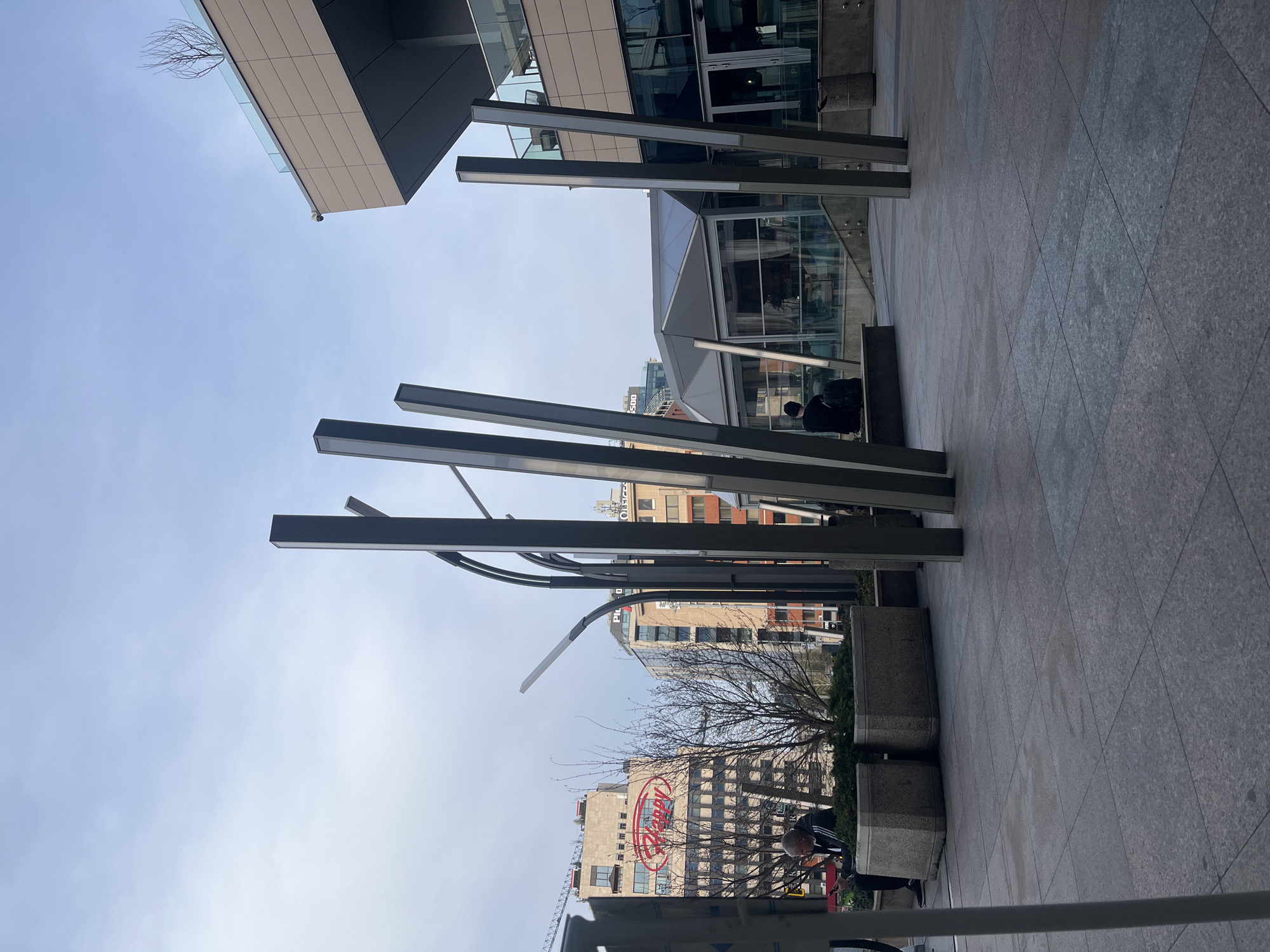 The image depicts an outdoor urban scene in Sofia, Bulgaria, characterized by modern architecture and street infrastructure. In the foreground are tall, rectangular metal light fixtures or decorative structures arranged in a semi-circular pattern. Behind these structures are raised concrete planters with small trees.
In the midground, various buildings are visible, including a large building with a prominent "Raffy" logo in red, suggesting a restaurant or similar establishment. Other buildings in the background exhibit a mix of architectural styles.
The sky is visible above the buildings and structures, appearing partly cloudy. The ground is paved with large, square stone or concrete slabs.
The scene contains people; one individual is seated on a bench near a building entrance, and another is standing near the planters.