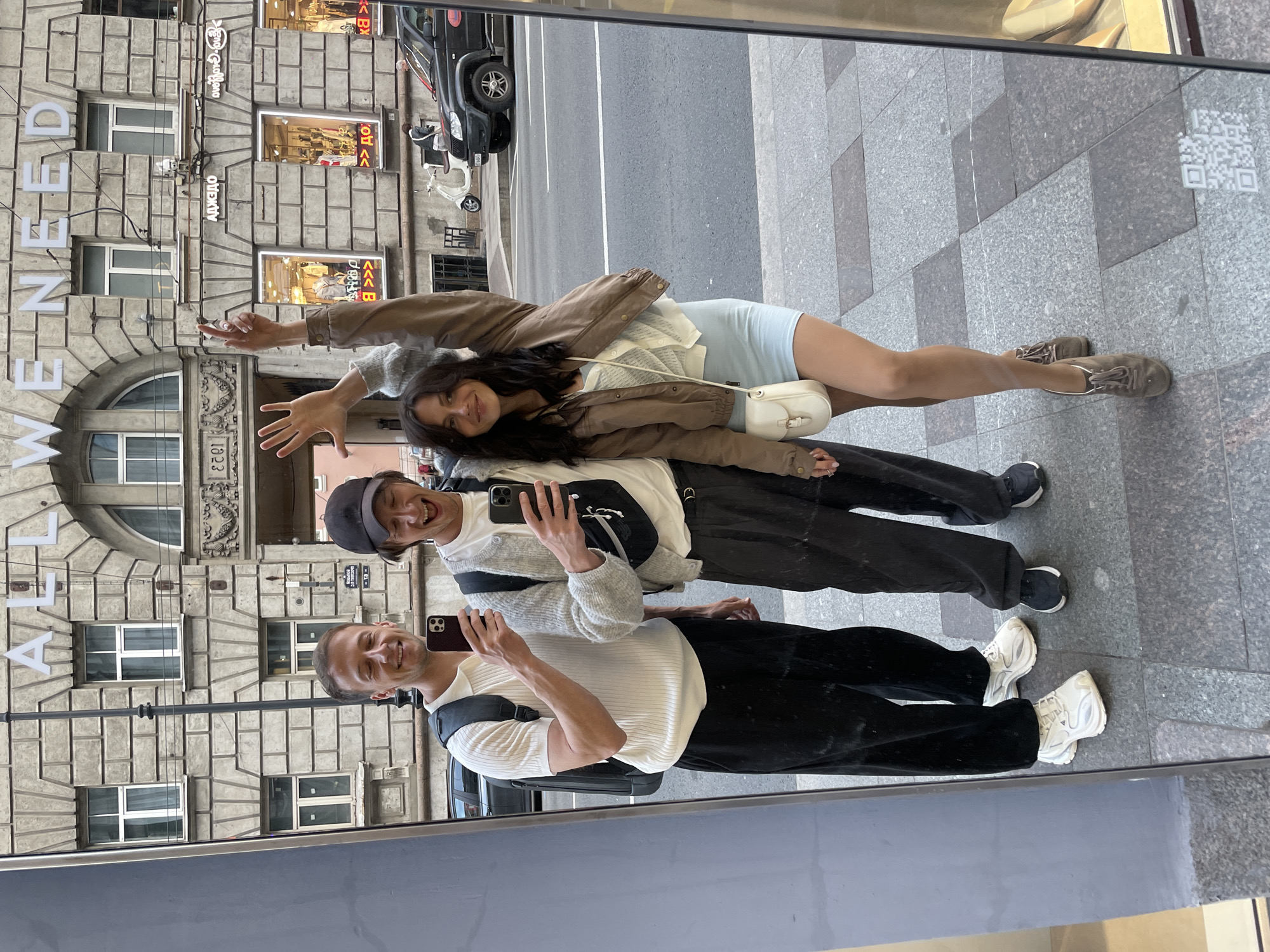 Three individuals are reflected in a large mirror, posing for a selfie outdoors in Saint Petersburg, Russia. The background consists of a building with ornate architectural details, including stone carvings and arched windows, with the words "ALL WE NEED" visible in large white lettering above. Street-level shops with displays of clothing are also visible through windows. A black SUV and a white scooter are parked on the street. The ground is paved with grey tiles.
On the left, a man wearing a white short-sleeved polo shirt and black trousers is holding a smartphone, taking the photo. He has short brown hair and is smiling with his eyes closed. A black backpack is visible on his back.
In the middle, another man wearing a white t-shirt, a light grey cardigan, and black trousers is also holding a smartphone and looking at the camera, laughing with his mouth open. He is wearing a black baseball cap. A black fanny pack is slung across his chest.
On the right, a woman with long dark hair is smiling at the camera, with her left arm raised and her right arm bent at the elbow, with her hand near her head. She is wearing a light blue mini-dress, a light brown jacket, and a white mesh cardigan. A small white crossbody bag is slung across her body. She is wearing brown lace-up shoes. The man on the left is wearing white sneakers, and the man in the middle is wearing dark sneakers.