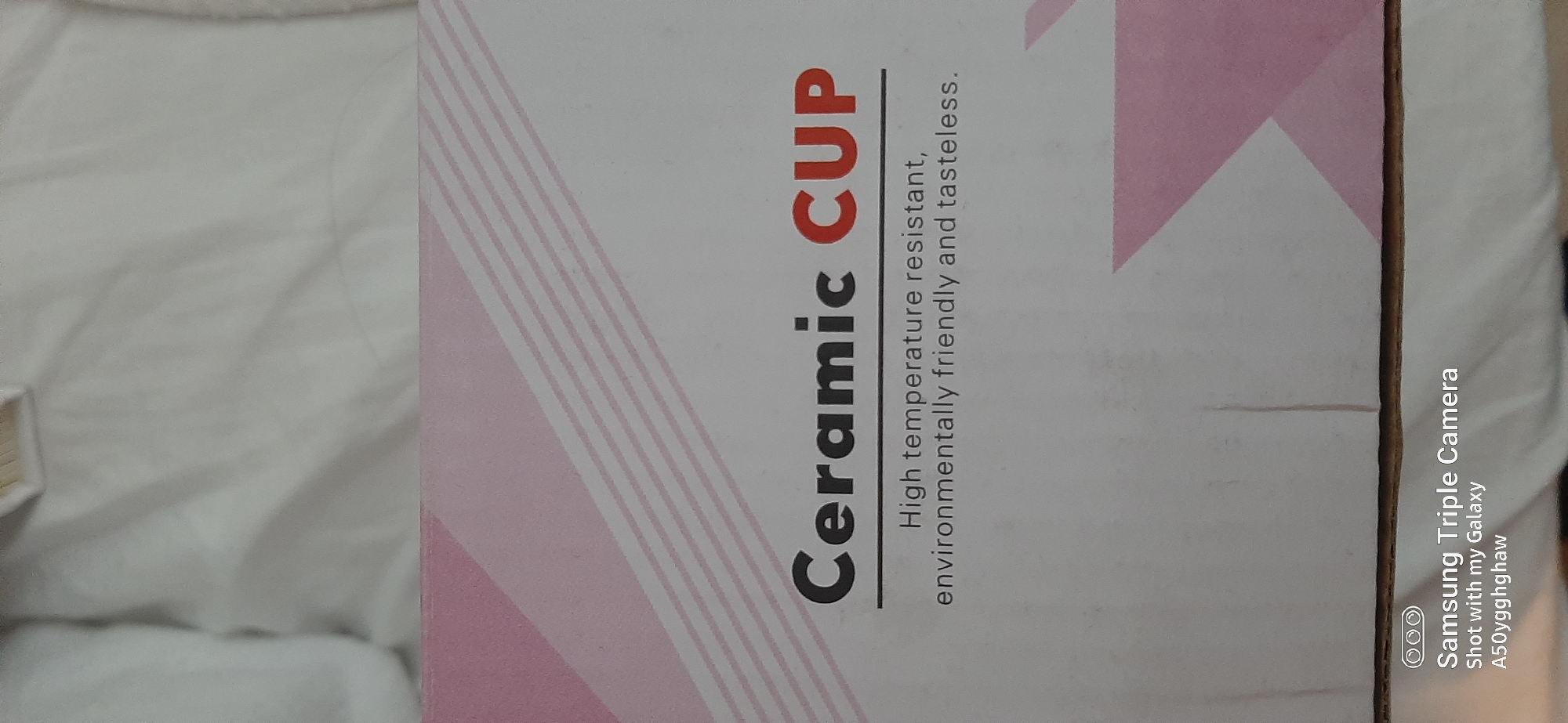 The image shows a close-up of a white cardboard box with pink diagonal stripes. Text on the box reads "Ceramic CUP" in large black and red letters, followed by "High temperature resistant, environmentally friendly and tasteless." in smaller black text. In the bottom left corner, there is a watermark that says "Samsung Triple Camera Shot with my Galaxy A50y9ghghaw". There is no indication of the city of Dubai, United Arab Emirates, in the image.