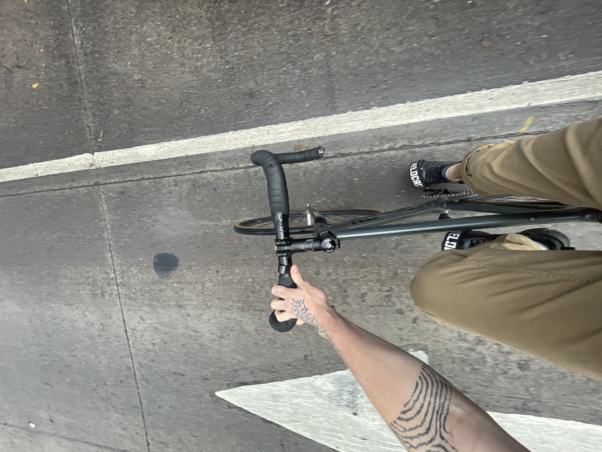 A high-angle, first-person perspective shot captures a person cycling on an asphalt road in Olivos, Argentina. The image is tilted, providing a view down towards the road, bicycle, and rider's limbs.
The rider is partially visible, wearing khaki-colored pants and black shoes with white soles. White text, "VELOCRA", is visible on the side of the right shoe. The rider's left arm, from the bicep to the hand, is prominently featured, covered in intricate black tattoos depicting wavy, topographic-like patterns. The hand firmly grips the black-taped drop handlebars of the bicycle.
The bicycle itself is partially shown, appearing to be a fixed-gear or single-speed model with a dark grey or black frame. The stem is visible, bearing the brand name "DEDA" on its cap. Closer to the rider's right foot, the brand "ALLCITY" can be seen on a part of the bicycle's frame or fork.
The setting is an asphalt road, marked by a clear, continuous white line running horizontally across the middle of the frame. A fainter, broken white line, possibly a crosswalk or edge marking, is visible in the lower left. The asphalt surface exhibits minor cracks, small debris, and a dark circular stain.
The activity depicted is cycling. The lighting suggests it is daytime, with an even distribution of light, indicating possibly an overcast sky or the absence of direct sunlight, as no harsh shadows are present.