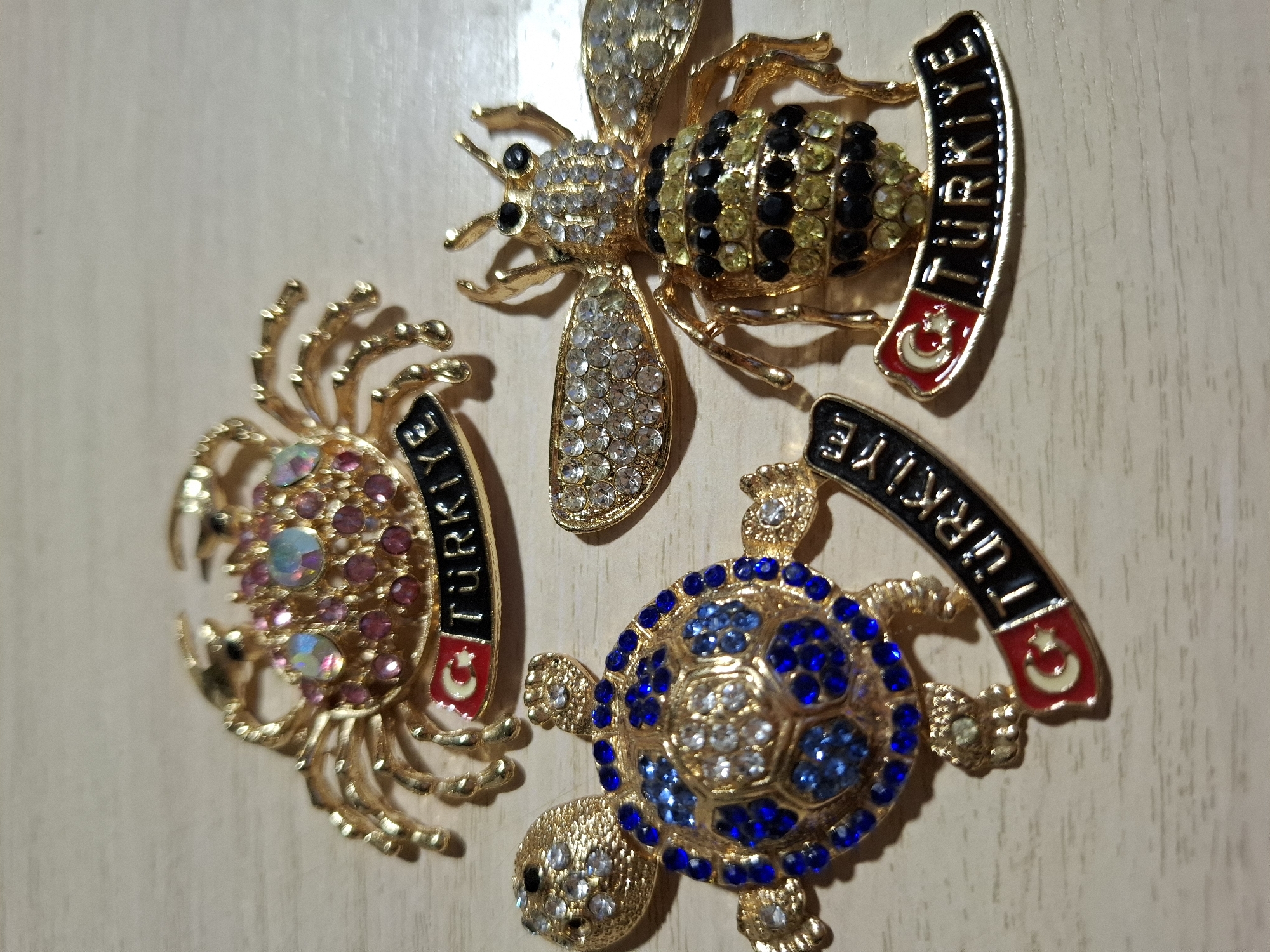Three gold-tone brooches are displayed on a light-colored wooden surface. From left to right, the brooches depict a crab, a turtle, and a bee. Each brooch is embellished with small, colorful rhinestones. The crab brooch has pink and iridescent rhinestones on its body and claws. It also features a black banner with the word "TURKEYE" in gold lettering, adorned with a Turkish flag. The turtle brooch is decorated with blue and clear rhinestones on its shell and head. It also has a banner with "TURKIYE" and a Turkish flag. The bee brooch has clear rhinestones on its head and wings, and black and yellow rhinestones on its body, arranged in stripes. It also displays a banner with "TURKIYE" and a Turkish flag. The wooden surface appears to be a table or countertop. There are no people or animals visible in the image. The scene is evenly lit, with soft shadows cast by the brooches. The location is not identifiable within the city of Tyumen, Russia.