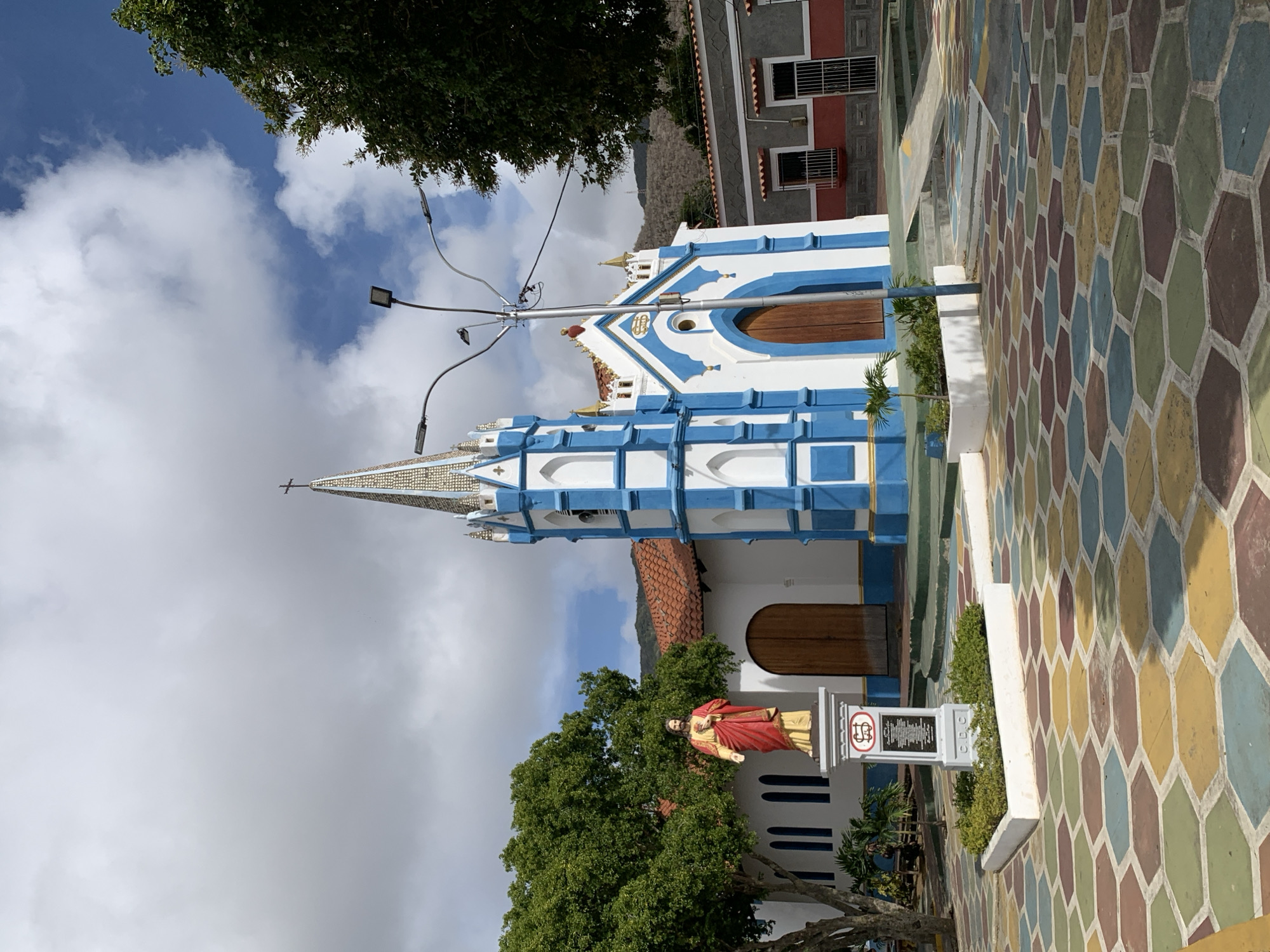 The image depicts the Church of Tacarigua in Tacarigua, Venezuela. The church is painted in white and blue, with a steeple topped by a cross. A statue stands in front of the church near a tree. The ground is covered in colorful, hexagon-shaped tiles. A street light is positioned in front of the church, with a building with red trim and windows in the background. The sky is partly cloudy.