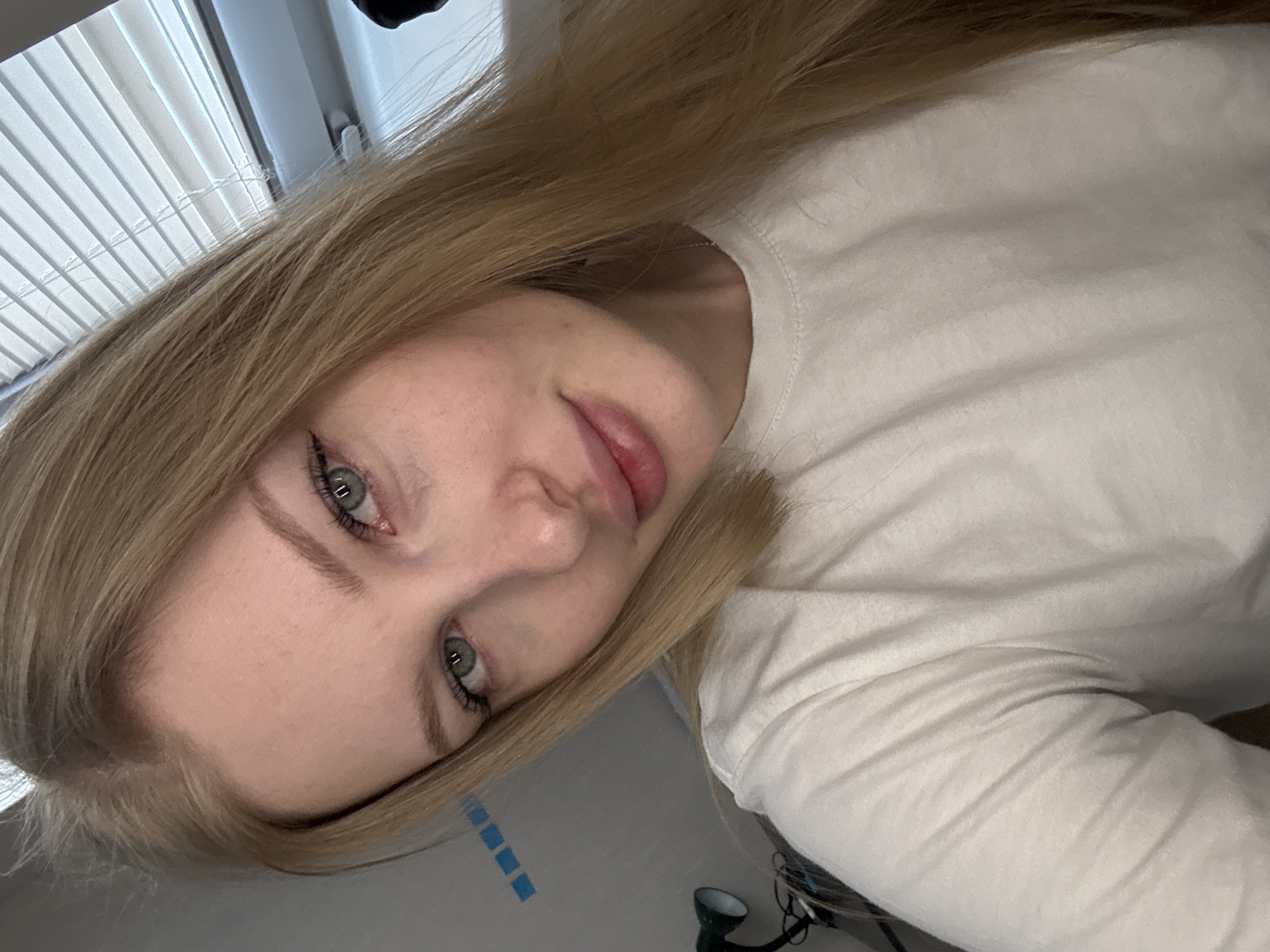 A young, fair-skinned woman with blonde hair and grey eyes is centered in the frame, angled slightly to the left. She is wearing a white long-sleeved shirt. The background includes white window blinds and a light grey wall with a strip of blue squares. A black desk lamp is partially visible at the bottom right corner. The location in Perm, Russia is not discernible from this image.