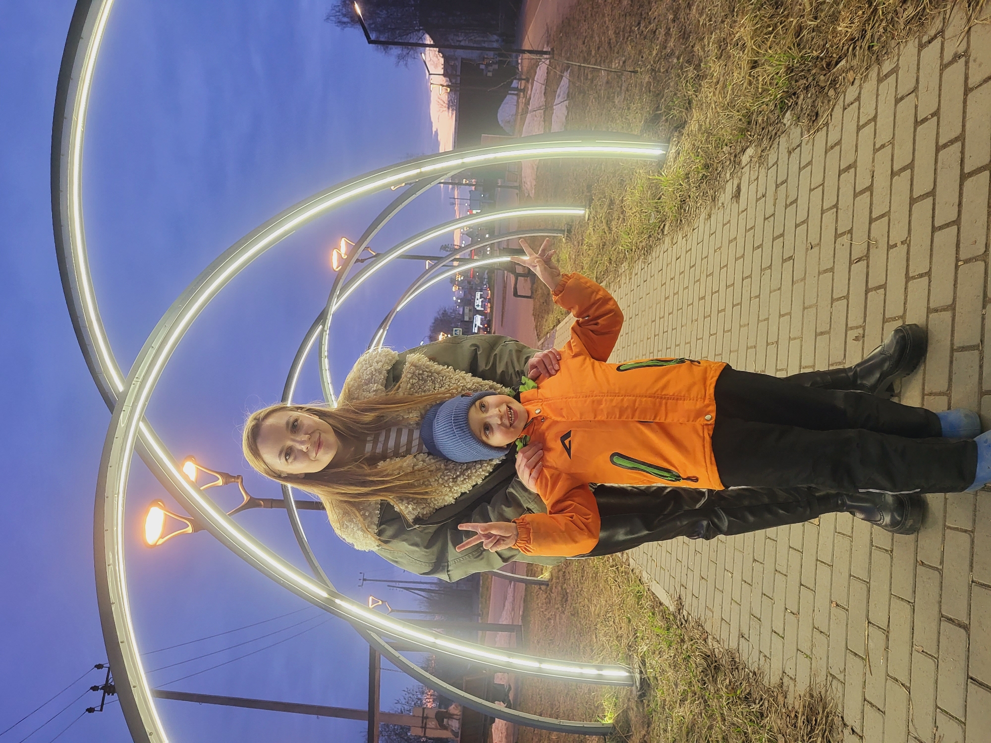 A woman and a child stand together on a brick walkway in Chaikovsky, Russia, under an illuminated public art installation. The woman has long brown hair and wears a green jacket with a tan shearling collar. The child, wearing a blue hat and an orange jacket, is making a peace sign with his right hand. The walkway is flanked by grass and the illuminated art installation, which consists of multiple glowing arches. The sky in the background is a twilight blue. Streetlights and buildings can be seen in the distance.
