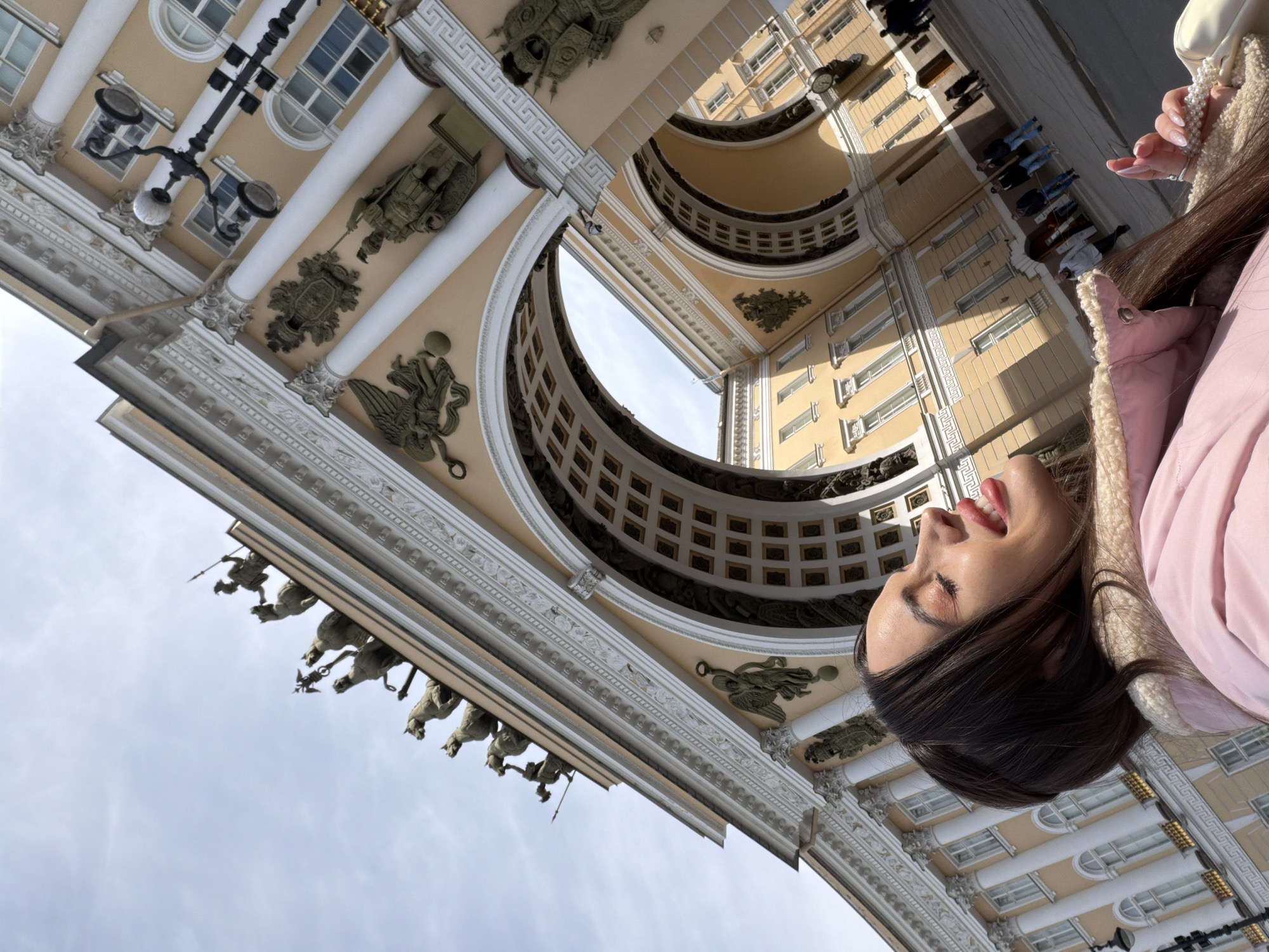 A low-angle shot of the General Staff Building in Saint Petersburg, Russia, shows the facade of the building with its archway and sculptures. A woman stands in the foreground, partially obscuring the view of the architecture. A group of people stand in the background.