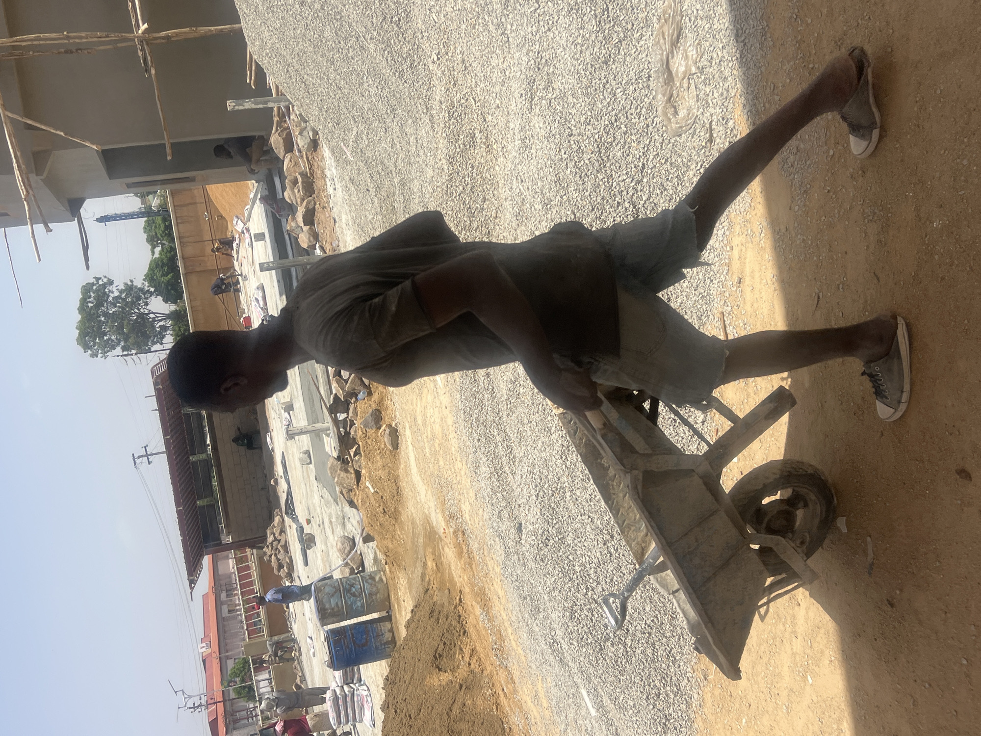 The scene is at a construction site in Barakin Lerawi, Nigeria. A man is pulling a wheelbarrow filled with gravel. The construction site has gravel spread across the ground and partially built structures are visible, including walls and wooden scaffolding. In the background are buildings, trees, and utility poles. Other construction materials like sacks are visible in the background. The man is wearing a t-shirt, shorts, and sneakers. He appears to be working.