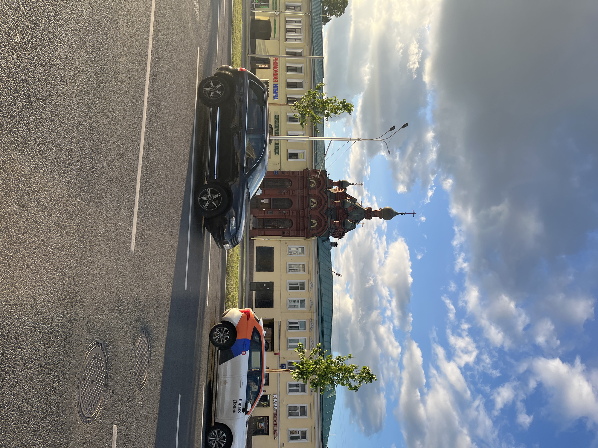 A street scene in an unknown city features two cars parked on the side of a road, adjacent to a row of buildings and under a partly cloudy sky. The first car, closest to the viewer, is a light-colored vehicle with an orange and blue accent. It has “Яндекс Драйв” and “Twenty Drive” written on its side. The second car is dark-colored. Behind the cars, there is a row of buildings with varied architectural styles, including a building with a red brick facade and ornate decorations. Street lamps stand beside the buildings, and trees with green leaves line the sidewalk. The sky is a mix of blue patches and white clouds, with some darker clouds in the upper portion of the frame. Manhole covers are visible on the road surface.