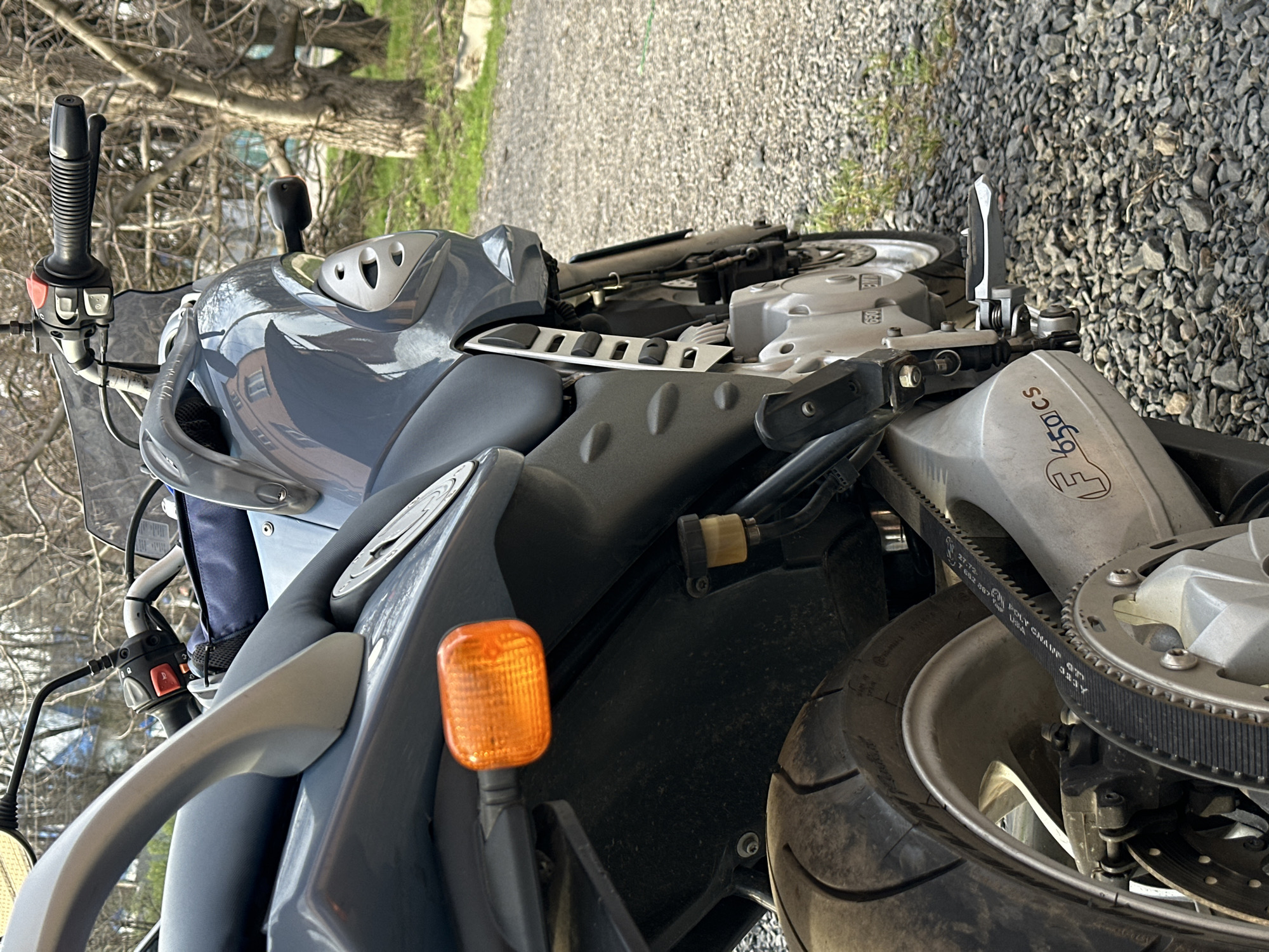 The image shows a gray BMW F650CS motorcycle parked on a gravel surface. The motorcycle is oriented so that the rear of the bike is facing towards the right side of the frame. The front of the motorcycle is visible on the left side of the image. The seat, part of the fuel tank, handlebars, turn signal, rear wheel, and belt drive are all visible. The rear wheel and the “F 650 CS” markings are visible. The background shows a blurry tree and what appears to be a paved area. No people are visible in the image. The location is not identifiable as being in Rostov-on-Don, Russia.
