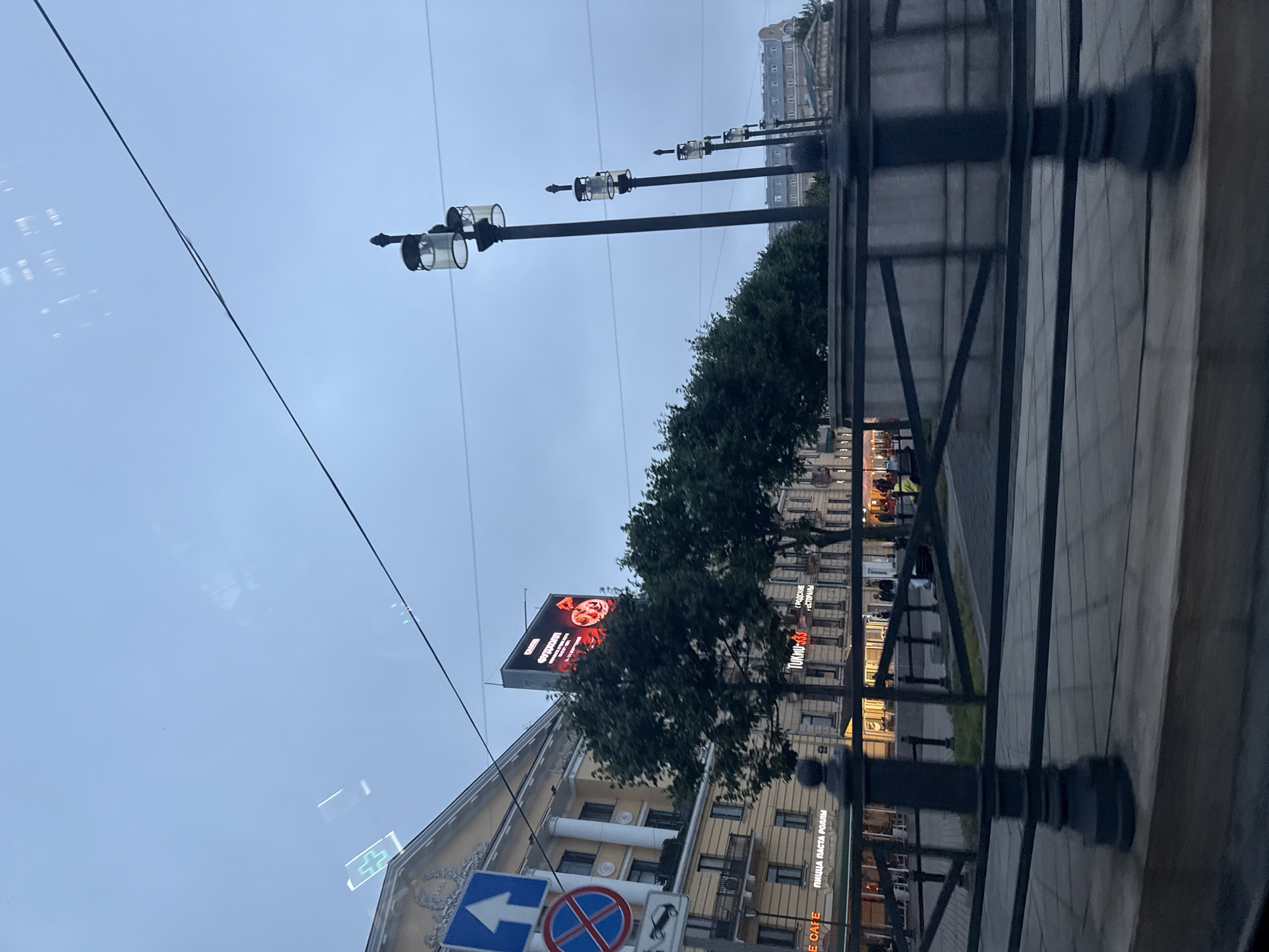 The image depicts a view of an urban street in Saint Petersburg, Russia, likely taken from a moving vehicle. The sky is overcast and a pale blue, suggesting early morning or evening.
In the foreground, a dark metal fence with decorative posts runs horizontally across the frame, partially obscuring the view of the street and sidewalk behind it. The fence is positioned over a grey stone embankment.
Further back, a sidewalk is visible, paved with light-colored tiles. A row of mature green trees forms a natural barrier between the sidewalk and the buildings.
Beyond the trees, a row of buildings lines the street. The building on the left is a light yellow, neoclassical structure with columns and balconies. A large digital billboard is mounted on this building, displaying text and an image that appears to be an advertisement, possibly for food. Traffic signs, including an upward-pointing arrow and a circular sign with a red cross, are visible on the left side of the building.
Several streetlights with glass enclosures are spaced along the sidewalk, adding to the urban ambiance. In the background, more buildings with pitched roofs are visible, characteristic of Saint Petersburg's architecture.
A few people can be seen on the sidewalk, dressed in casual clothing. They appear to be walking or standing, their presence adding a sense of activity to the scene. The lighting is subdued, and there are reflections on the glass from which the photo was taken, suggesting it was captured through a window. The overall impression is one of a typical urban environment in Saint Petersburg during twilight hours.