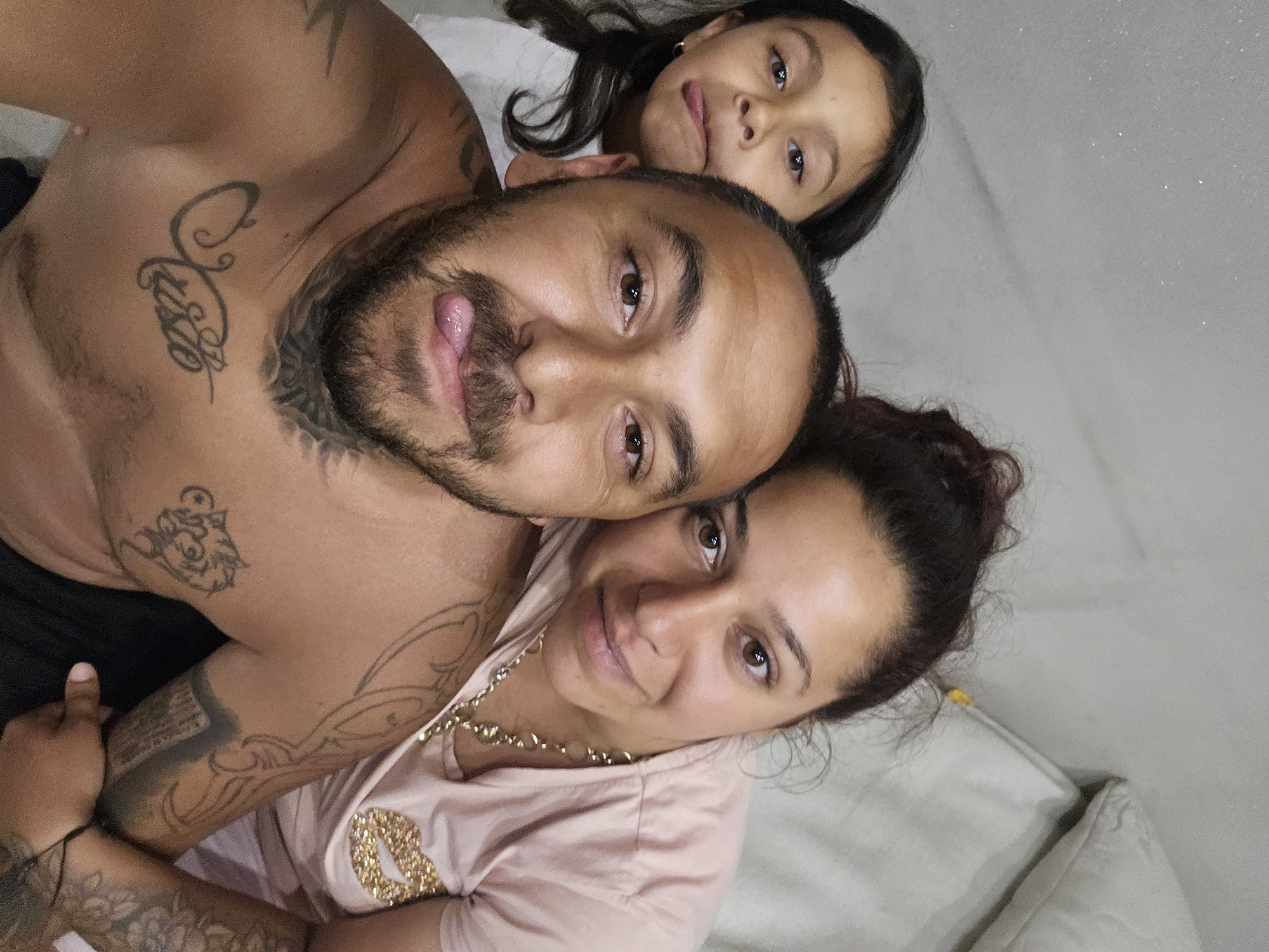 A man, woman, and girl are taking a selfie. The man is in the foreground, shirtless, with tattoos on his chest and arms, sticking his tongue out. The woman is to his right, wearing a pink shirt with a gold lip print design and a gold chain. She is smiling. The girl is to the man's left and behind him, wearing a white shirt, and also sticking her tongue out. The background appears to be an indoor setting with a light-colored wall and what looks like a pillow. The image does not contain any information that would indicate its location in Sofia, Bulgaria.