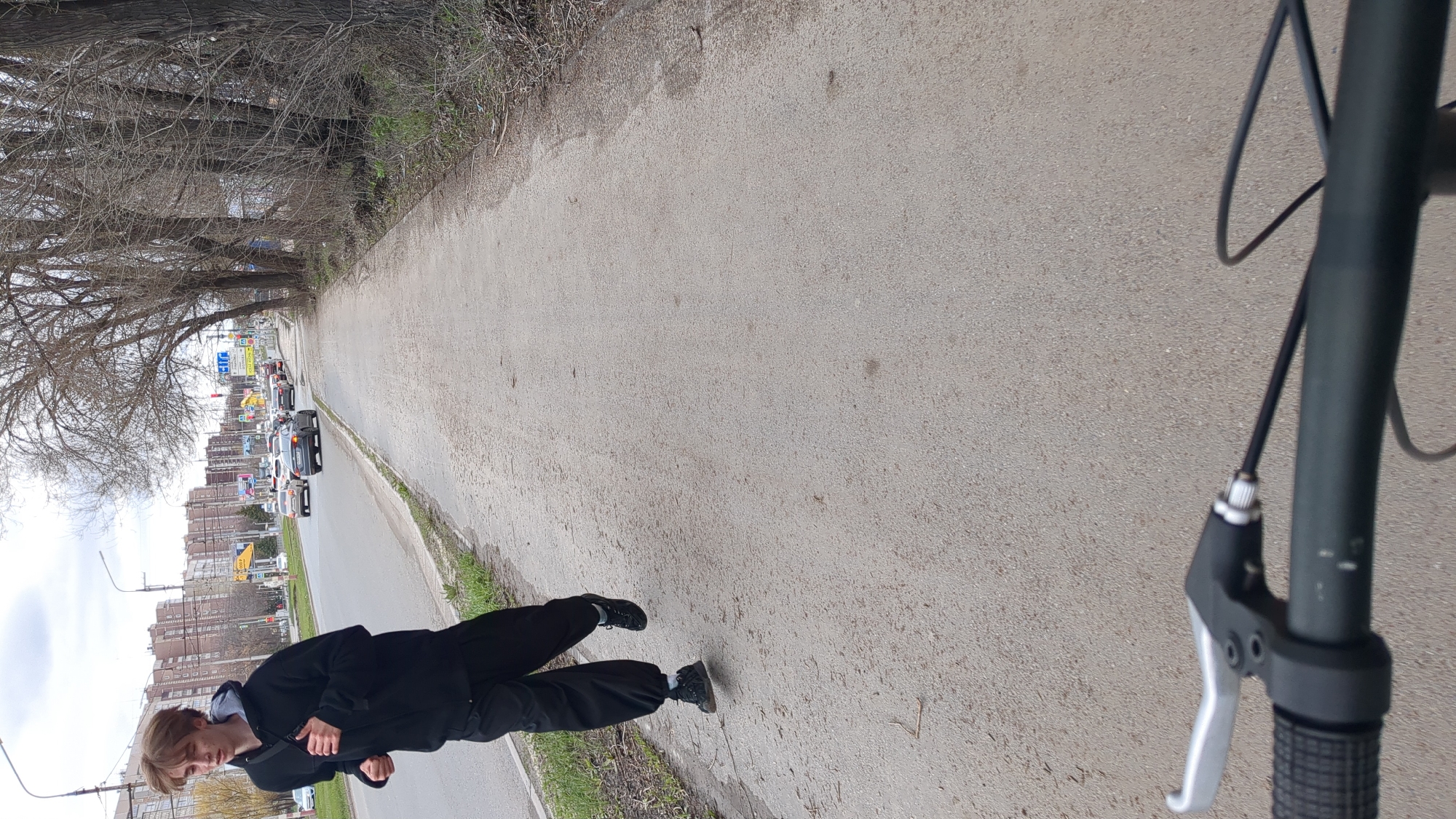 The image shows a person standing on a paved sidewalk next to a road. The person is wearing a dark outfit and appears to be looking at a phone or small device. In the foreground, there is a section of a bicycle handlebar. The road has several cars in it and is lined with trees on the sidewalk side. Buildings can be seen in the background of the image, and signs with street names are visible. Based on the urban environment, buildings, signs and street configuration, it is likely that the location is in the city of Ulyanovsk, Russia.