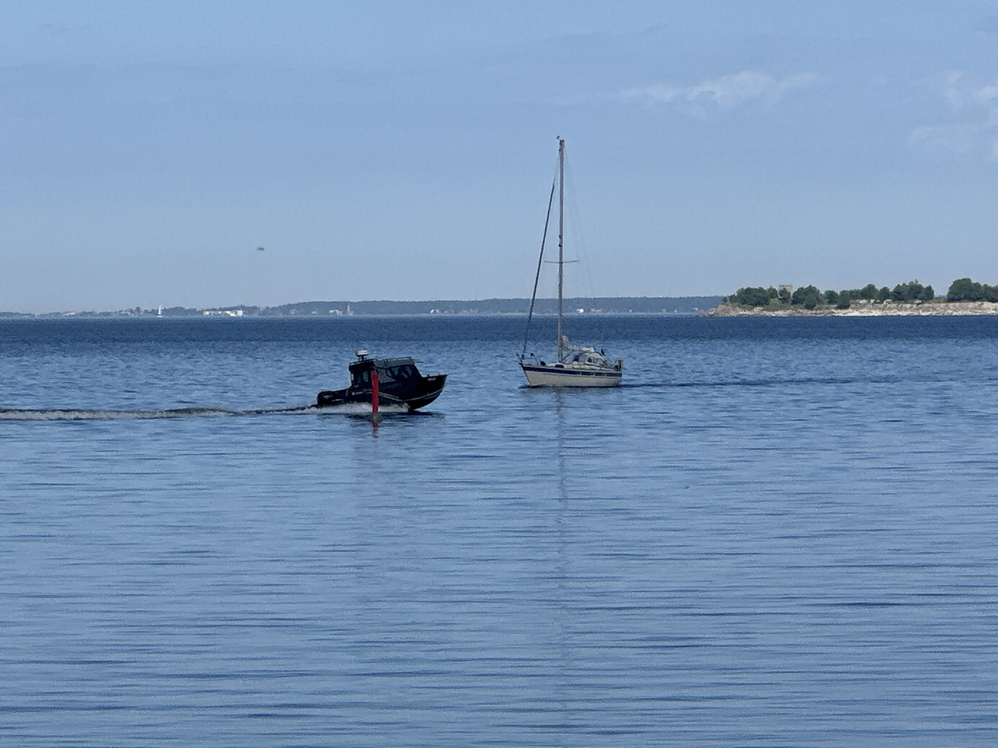 The image depicts a serene maritime scene under clear daylight, likely within Tallinn Bay, Estonia. The foreground and midground are dominated by calm, light blue water exhibiting subtle horizontal ripples, suggesting minimal wind. Two boats are visible: on the left, a dark-colored motorboat with a small cabin is actively moving from left to right, leaving a distinct white V-shaped wake behind it. A prominent red cylindrical marker or buoy is positioned vertically in the water near the motorboat's bow. To the right, a stationary light-colored sailboat with a single tall mast and a white hull with a darker stripe is present. The horizon reveals two distinct landmasses. The distant land from the left to the center features a low-lying, urbanized coastline with various buildings and structures, including a discernible white lighthouse or tall tower on the far left. To the right, a closer, tree-covered peninsula or island juts into the water, characterized by green deciduous trees and a small, light-colored structure nestled among them. The sky above is a pale blue with only minimal, faint white cloud formations. No individuals are visibly present on either boat or the distant land.