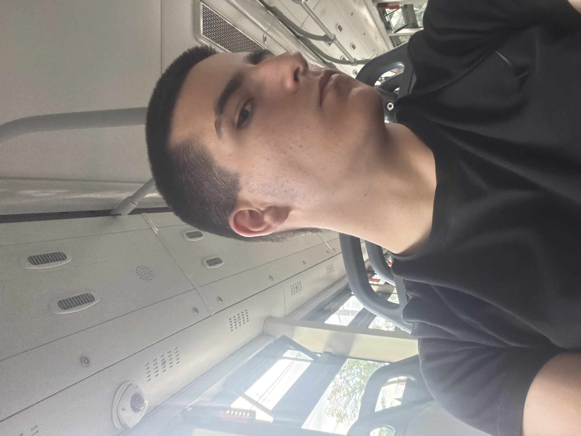 The image shows a young person with short dark hair and dark eyebrows, wearing a black t-shirt, sitting inside a bus. The person is looking to their right, with their face partially visible. The background shows the interior of a bus, including seats, handrails, windows, and the ceiling with vents and a camera. The image is taken from a close-up perspective, focusing on the person and their immediate surroundings. The bus appears to be in motion, as indicated by the blur of the exterior through the windows, which show trees and some buildings, suggesting an urban or suburban environment. The location of Kubratovo, Bulgaria is not visually evident from the image itself, but it is provided as context.