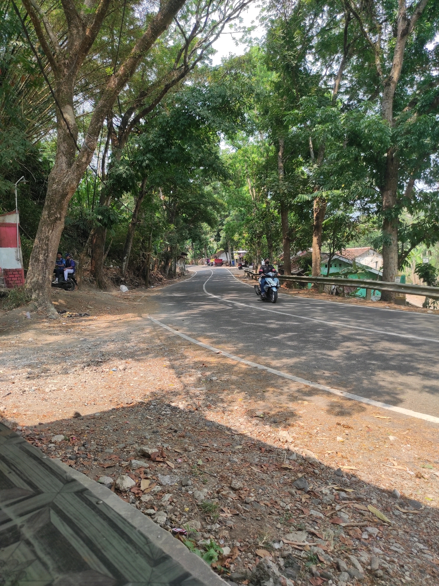 The image depicts a rural road in Mekarwangi, Indonesia, under a canopy of lush green trees. A paved road curves through the scene, with a white line marking the lane. On the left side of the road, two individuals are seated on a black motorcycle parked on a dirt and gravel shoulder. Behind them, a red and white structure is partially visible. Further down the road, another person is riding a light blue scooter, heading away from the viewer. The right side of the road is bordered by a metal guardrail, beyond which are houses with green and white exteriors, interspersed with trees and vegetation. The ground on the shoulder is covered with scattered rocks, dried leaves, and some debris. The overall impression is a sunny day in a tropical, semi-urban or rural setting.