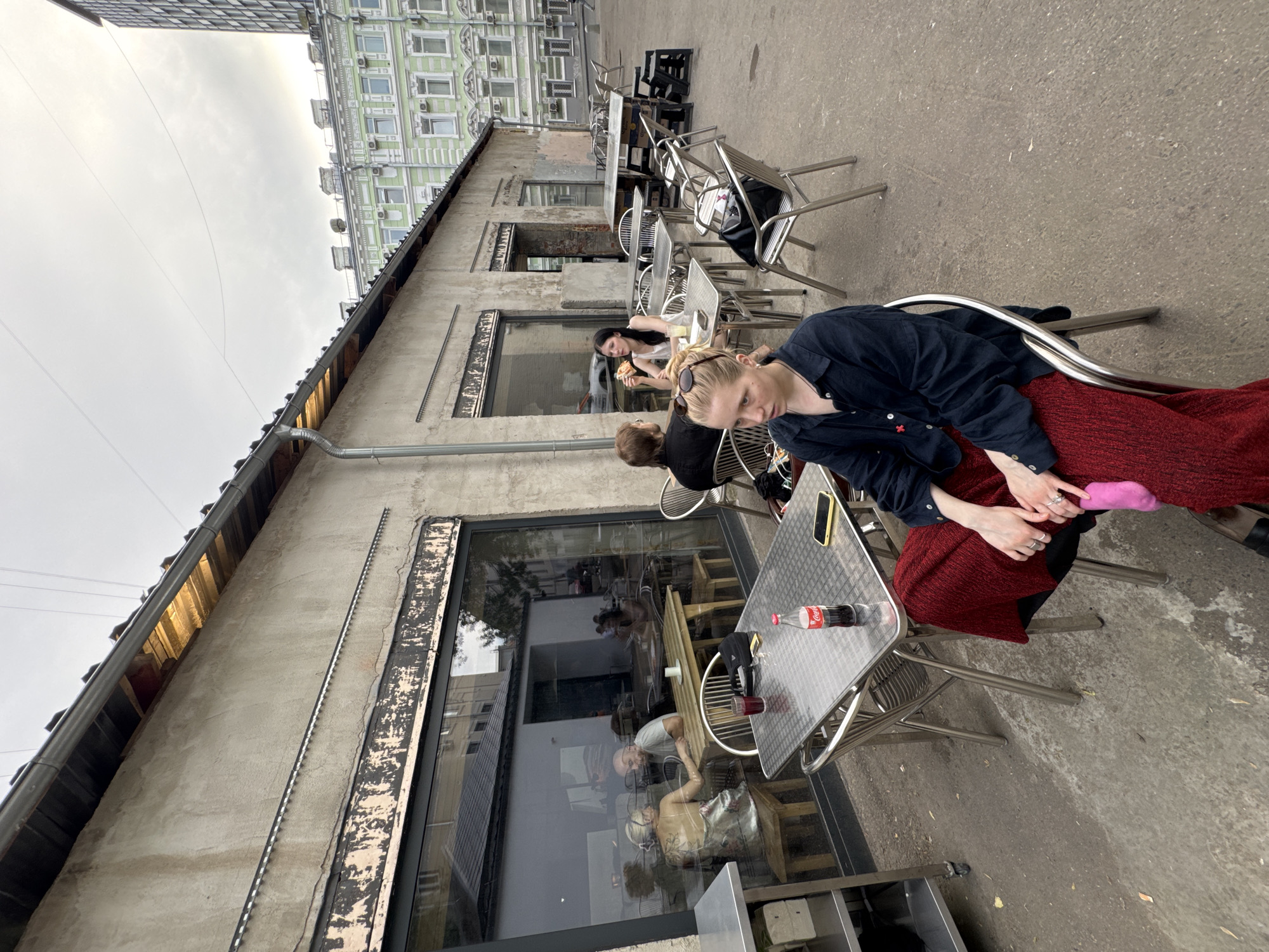 The image depicts an outdoor dining area in Moscow, Russia. Three people are visible seated at metal tables along the exterior of a long, weathered building with large windows. A woman in a dark blue shirt and red skirt sits at the nearest table, her hands clasped in her lap. A bottle of Coca-Cola and a cell phone are on the table. Another person, wearing black, is seated next to her. At the next table, a third woman is eating. The building has a gray, weathered facade. Across from the building is a green and white building. The dining area is located on a paved surface. The sky is overcast.