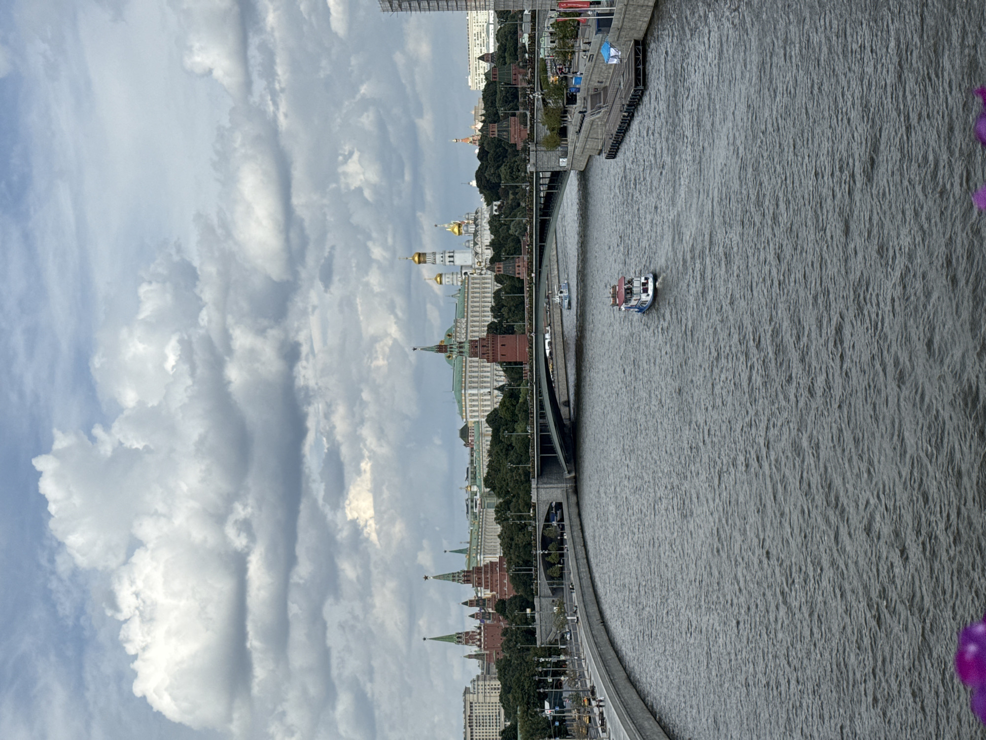 This is a medium shot, taken from an elevated perspective, of the Moskva River flowing through Moscow, Russia. The river dominates the foreground, its surface rippled by a gentle current. Two small white boats with red roofs are visible on the water. The left bank of the river features a curved embankment and a wide pedestrian walkway lined with streetlights. Beyond the walkway, a multi-lane road runs parallel to the river, with a few cars visible.
In the midground, a large bridge with stone arches spans the river. Behind the bridge, the Moscow Kremlin complex is prominently displayed. This iconic landmark features several distinct red-brick towers topped with green spires, some adorned with gold stars. Among these are the Kremlin's famous cathedrals, with their golden domes glinting under the cloudy sky. The Kremlin is surrounded by lush green trees.
The background is a cityscape with various buildings, including a large white building with a green roof and other structures of varying architectural styles. The sky above is filled with dramatic, grey, and white clouds. In the very bottom foreground, out of focus, are two purple flower petals. The overall lighting suggests an overcast day.