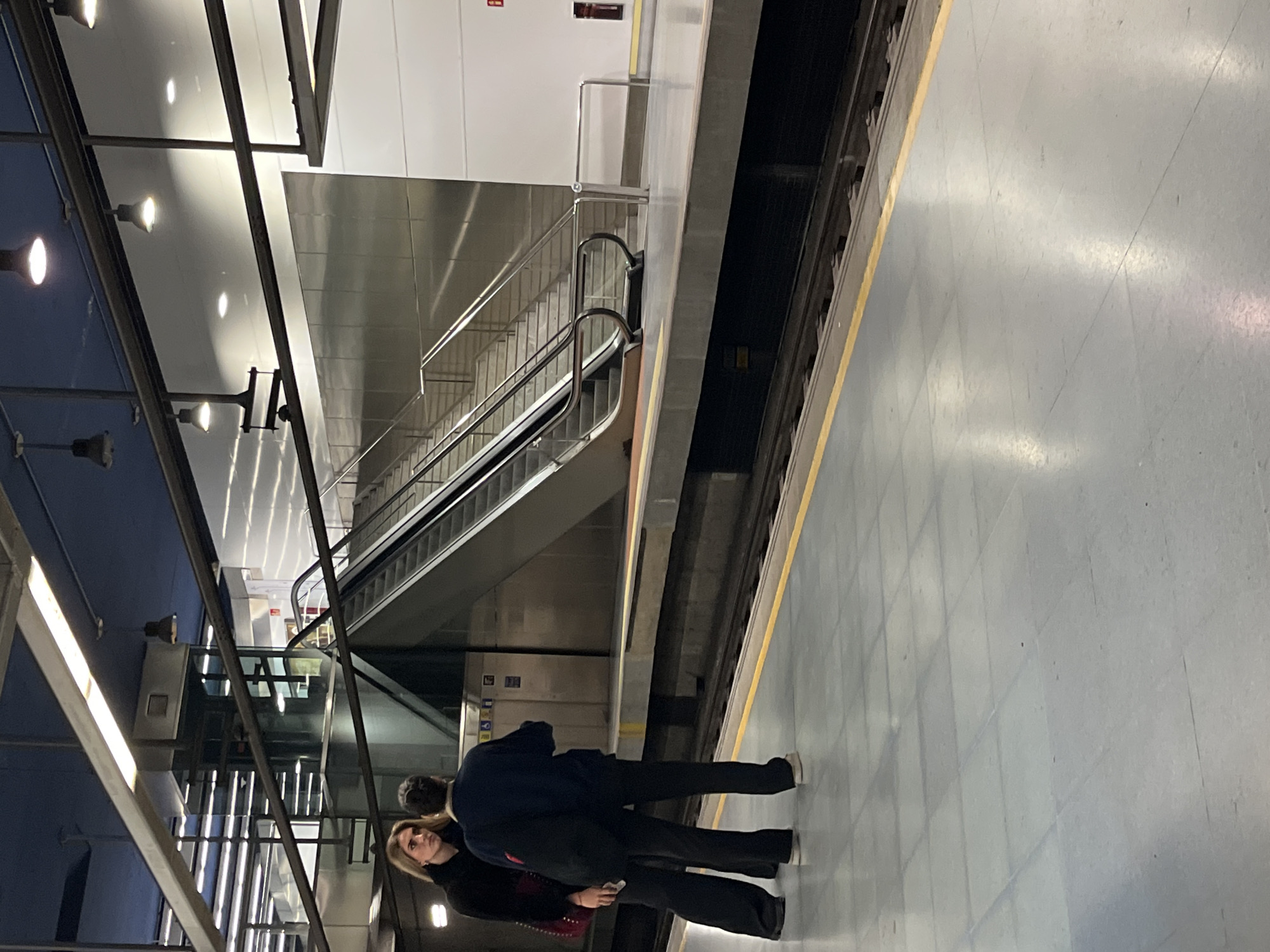 The image depicts a scene in a subway station platform, likely in València, Spain. The platform is lined with a yellow safety strip and has a gray tiled floor with rectangular patterns. In the foreground, two people stand, one with blonde hair and wearing a black jacket and the other person wearing a black shirt. Adjacent to the platform is an escalator, which is located beneath the station's lighting. The lighting consists of a series of overhead lights and metal framework. The background is composed of a wall with stainless steel panels.