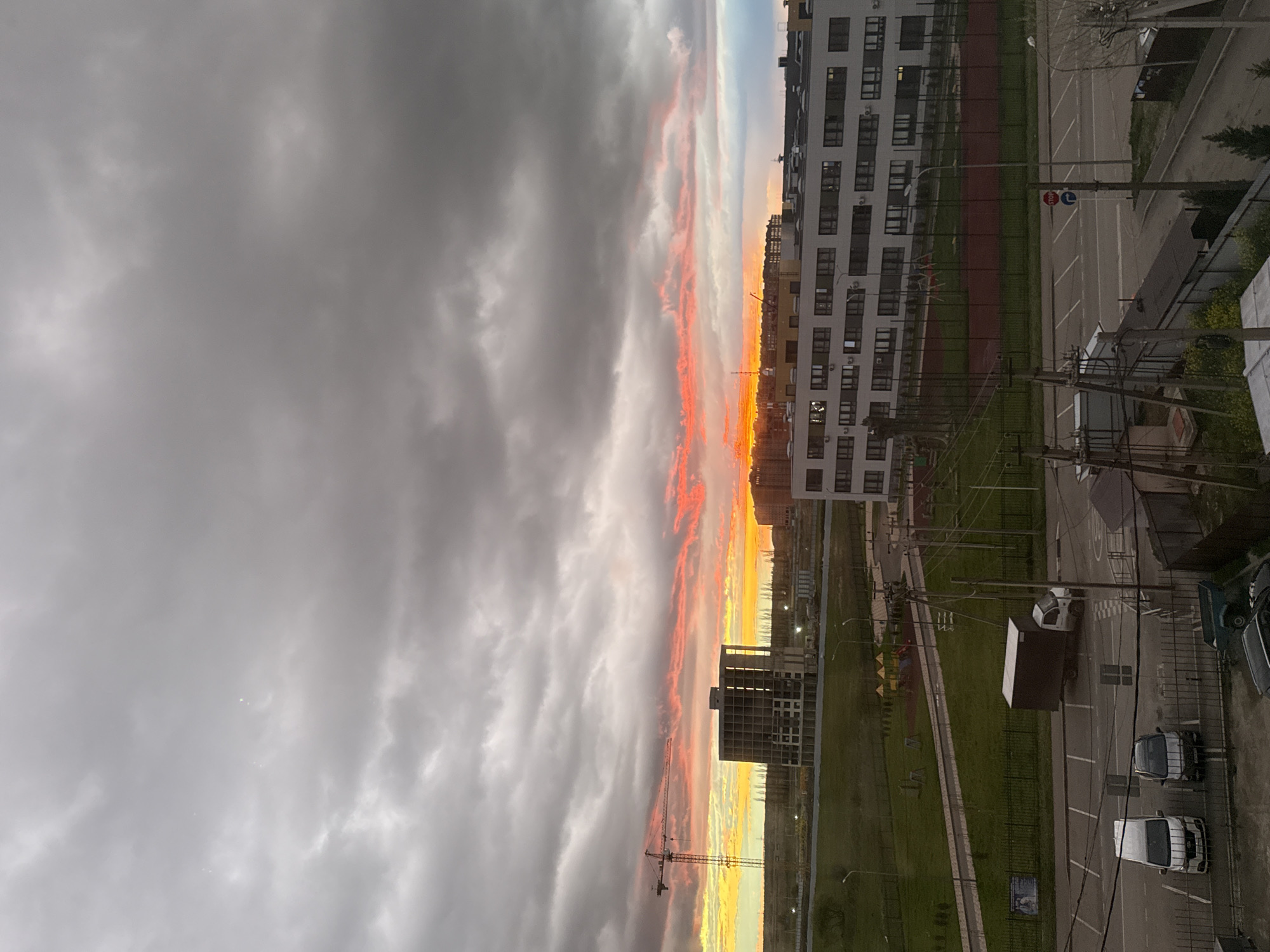 The image shows a twilight cityscape with a colorful sunset. In the foreground and to the right, there are buildings, a parking lot with several parked vehicles, and green spaces. In the distance, the sky is filled with gray clouds on the left, transitioning to orange and yellow hues where the sun is setting on the right. A construction crane stands amidst the buildings, silhouetted against the brightly colored sky.