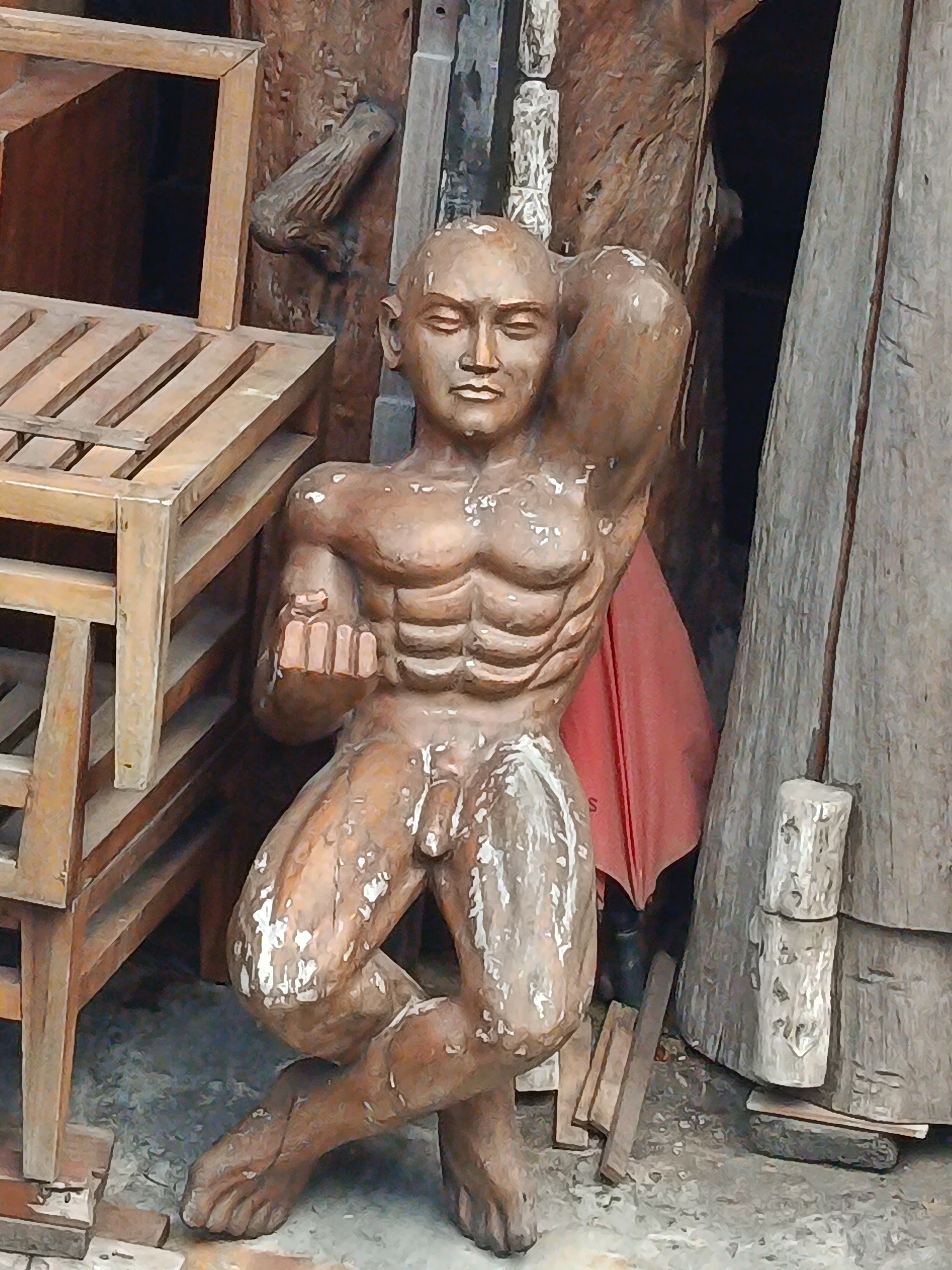 A weathered wooden sculpture of a muscular, nude male figure kneels on a gray, gritty surface. The sculpture has a bald head, closed eyes, and a determined expression. Its left arm is bent at the elbow, hand held up near its face, while its right arm is raised over its head, its hand resting on its bicep. The figure's torso is prominently defined with sculpted abdominal muscles. Its right leg is bent and tucked underneath its body, while its left leg is extended forward. The sculpture's surface is textured and shows signs of age and wear, with white paint peeling off in places.
The sculpture is situated in an outdoor setting, surrounded by a variety of wooden objects. To the left of the sculpture is a portion of a wooden rack or shelf unit made of light-colored wood with horizontal slats. Behind the sculpture, tall, dark wooden structures and a bright red fabric draped over something are visible. To the right, a large, rough-hewn wooden post or beam stands vertically, and a smaller wooden block with carved markings rests against it. The ground is covered in loose gravel and debris.
The overall impression is of an art installation or display in an outdoor market or workshop in Kuta, Indonesia, possibly a craft area given the abundance of wood and handmade items.
