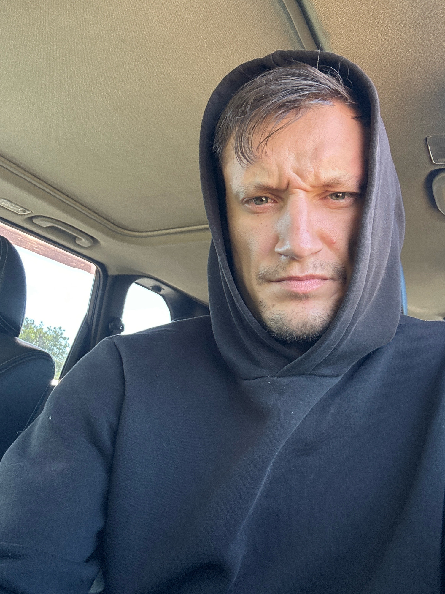 In Makiv, Ukraine, a close-up selfie taken from inside a car shows a man wearing a black hoodie with the hood pulled up, obscuring his head and hair. He is looking directly at the camera with a furrowed brow and a stern expression, with his eyes slightly narrowed and his mouth closed. The man has a light beard and stubble. The interior of the car is visible, with the beige headliner and a portion of the sunroof. Through the car window, trees and foliage are visible against a bright sky, indicating an outdoor setting. The lighting suggests it is daytime.