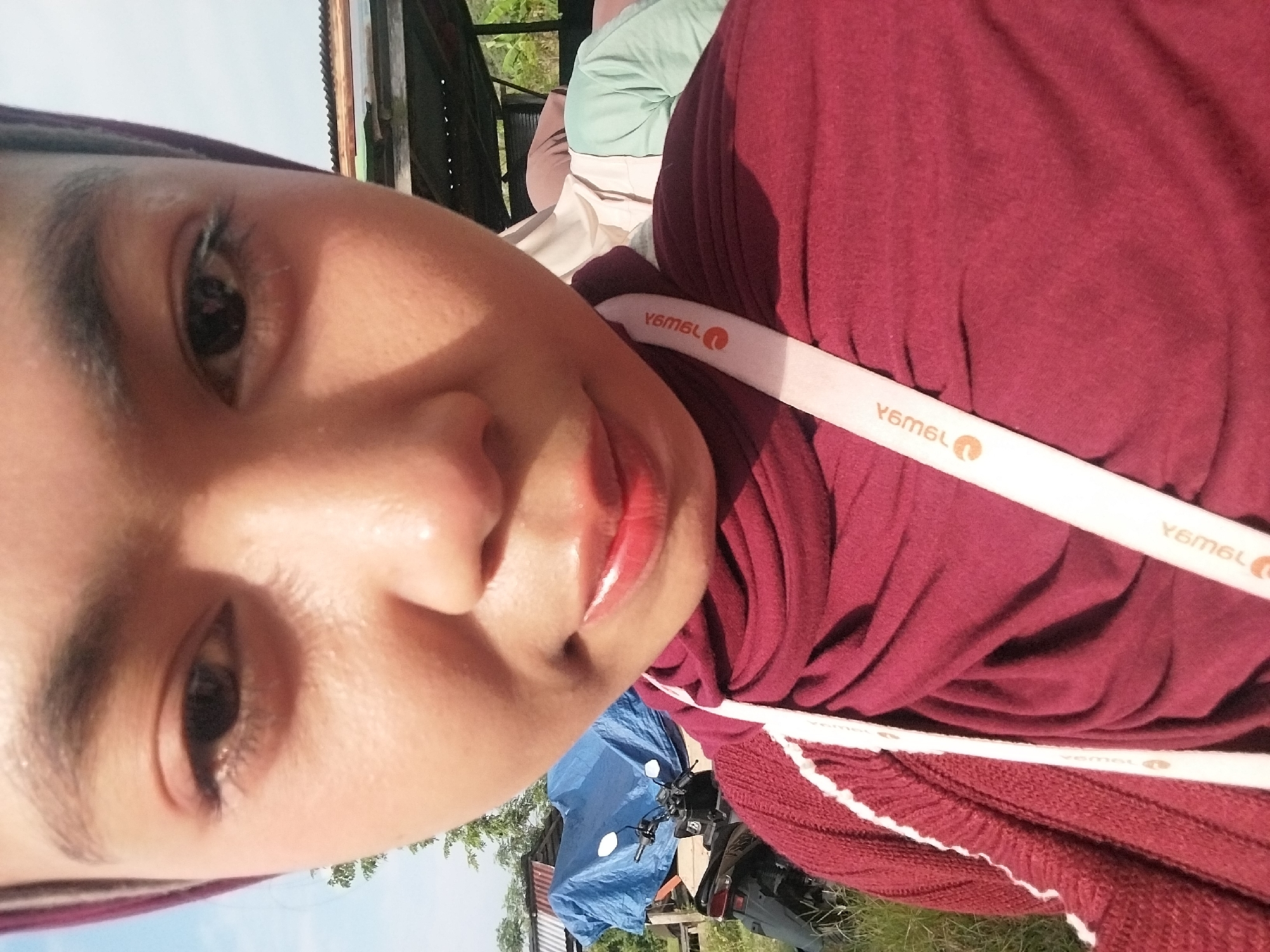 A close-up, upward-facing selfie shows a young woman wearing a maroon hijab and a maroon sweater. A white lanyard with orange text is draped over her chest. Her right eye is clearly visible, while her left eye is mostly obscured. Her lips are painted red. In the background, a blue tarp covers an object, and a motorcycle is parked beside it. A rustic wooden structure with a corrugated metal roof is visible behind the tarp, and some greenery is present in the distance. The scene appears to be outdoors, possibly in a rural or semi-rural area. The lighting suggests it is daytime. The location is identified as Turumame, Indonesia.