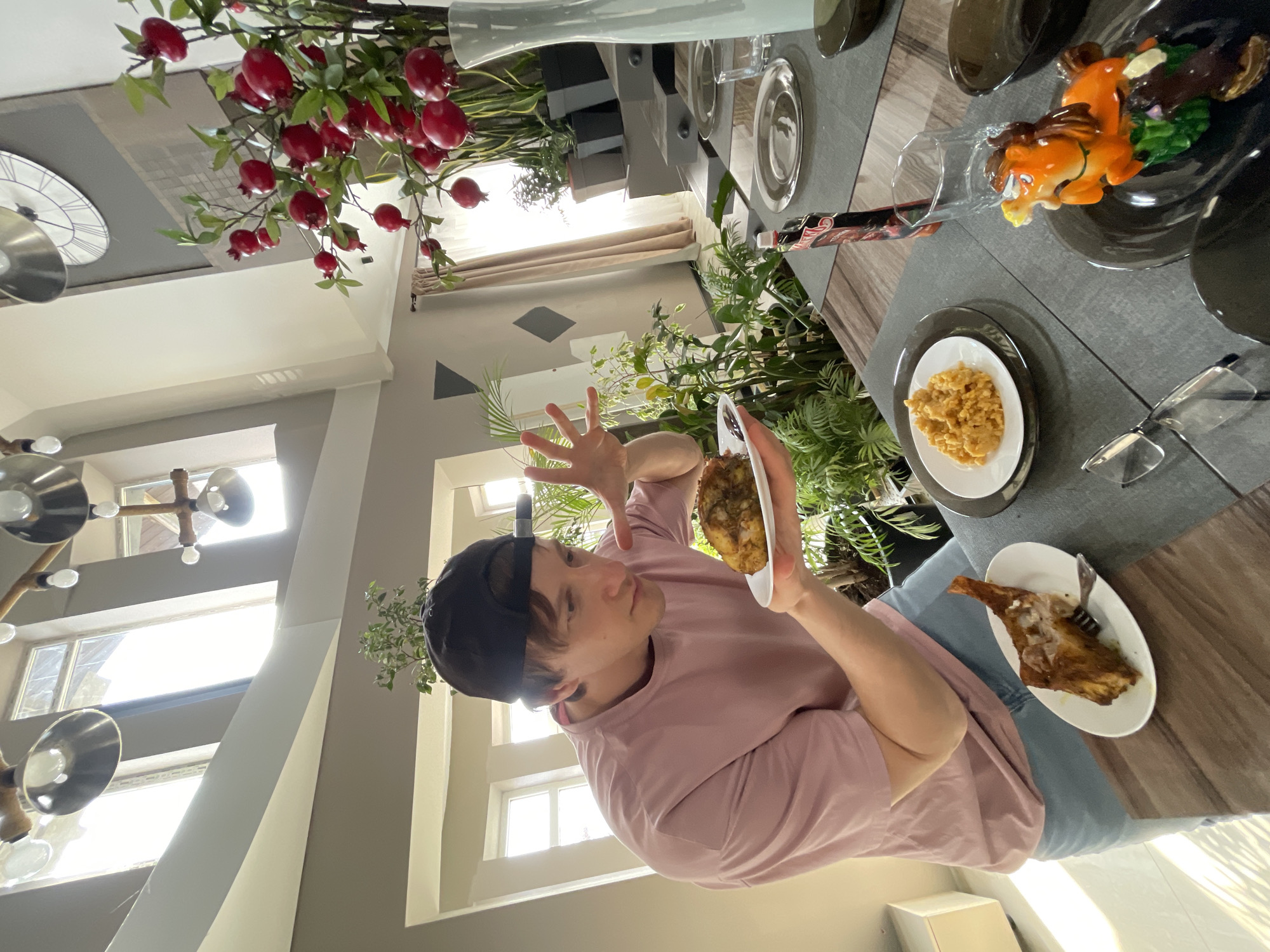 A young person in a pink shirt and backwards black baseball cap holds a plate of food. The person stands near a dining table with other plates of food, including one with a ceramic horse figurine. There are plants and decorative pomegranate branches on the table and in the background. A window is visible behind the person, suggesting the setting is indoors. The image does not provide enough information to confirm the location in Kursk, Russia.