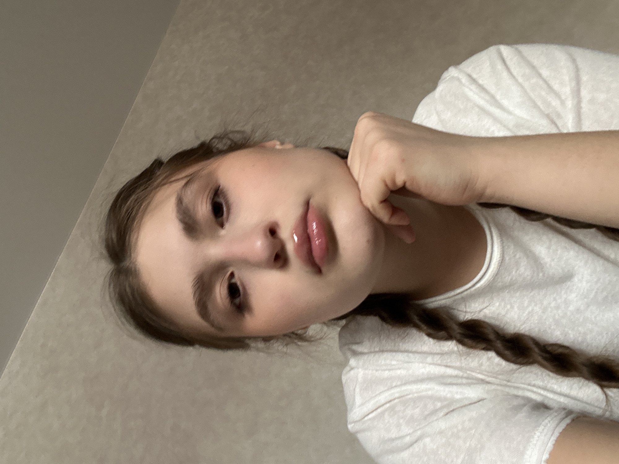 The image displays a head and upper torso portrait of a female subject, estimated to be in her late teens or early twenties, photographed indoors. She possesses light skin, dark eyes, and dark brown hair styled into at least two visible braids, with one prominent braid resting over her left shoulder on her chest. Her lips are full and appear to be coated with a clear or light pink gloss. She is wearing a light-colored, possibly white, short-sleeved t-shirt with a textured fabric. Her right hand is positioned beneath her chin, with her fingers lightly fisted, appearing to support her head. Her gaze is directed forward, towards the camera. The background consists of a plain, light beige or light brown textured wall, indicative of an indoor residential or private setting. A ceiling line is visible in the upper right quadrant. No specific environmental markers are present to identify a precise location within the city of Sarapul, Russia.
