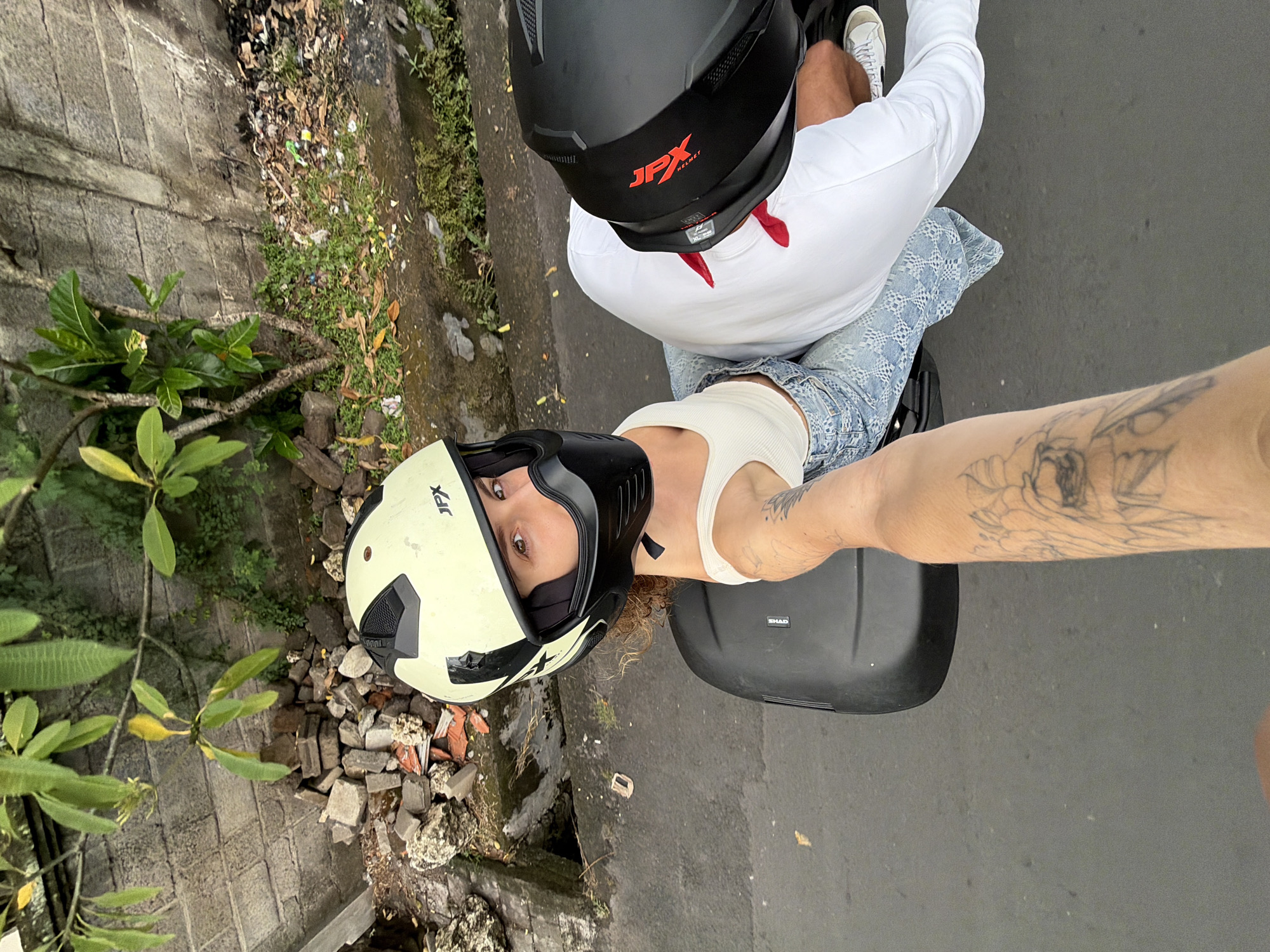 In North Kuta, Indonesia, two individuals are seated on a scooter, viewed from a first-person perspective. The person in the foreground, wearing a light-colored helmet with "JPX" branding and a black face mask, is looking towards the camera. They have curly hair and are wearing a white tank top and patterned denim jeans. Their left arm, adorned with tattoos, extends forward, suggesting they are holding the camera or a phone. Behind them, the second individual wears a black helmet with red "JPX" branding and a white long-sleeved shirt. They are also wearing shorts and white sneakers. The scooter has a black storage compartment. The environment shows a roadside with a brick wall, some greenery, loose bricks, and debris on the ground, indicative of an urban or semi-urban setting. The road surface is asphalt.
