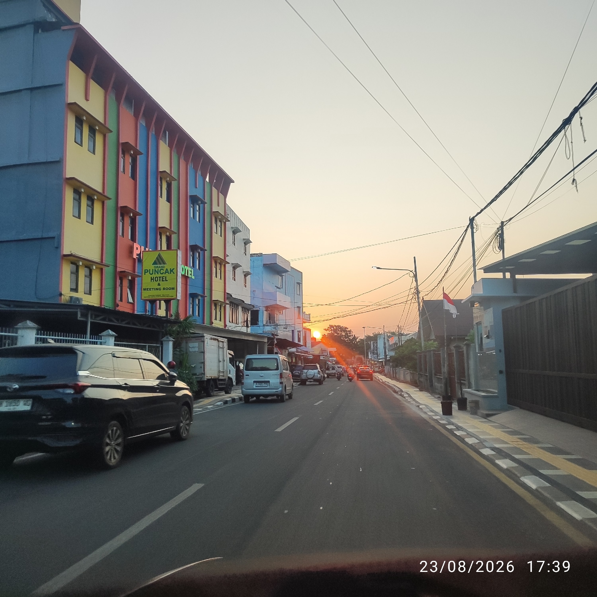 The image depicts a street scene in Tanjung Pandan, Indonesia, during the late afternoon or early evening, as indicated by the sunset. The foreground is dominated by the asphalt road and a portion of a vehicle, likely a car, seen from the rear passenger side. A white dashed line marks the lane. The road has a concrete gutter and a sidewalk with yellow and grey patterned tiles on the right.
On the left side of the street, a brightly colored, multi-story building stands out. Its facade is a vibrant mix of blue, yellow, red, and green panels, with visible windows. A prominent green sign with yellow text reads "GRAND PUNDAK HOTEL & MEETING ROOM." Adjacent to this building, other structures are visible, including a white truck and a lighter-colored van.
Further down the road, traffic is present, including several cars, a motorcycle, and another van. The setting sun is visible in the distance, casting a warm glow and lens flare effects on the road and surrounding buildings. Power lines crisscross the sky. On the right side of the street, a modern-looking building with a dark, slatted fence and a light-colored roof is present. An Indonesian flag, red and white, is planted on a pole in front of a residential-style building further down the street. The timestamp "23/08/2026 17:39" is overlaid on the bottom right corner of the image.