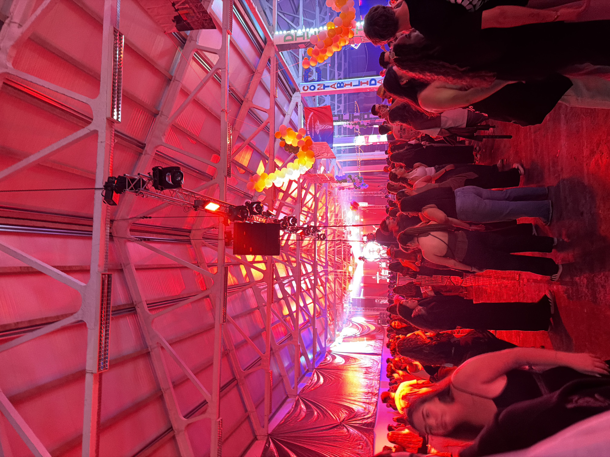 Correctly oriented, the image captures a vibrant indoor event, likely a party or concert, in a large venue, possibly in Bragança, Portugal. The scene is dominated by striking red lighting, bathing the entire space in a rich, dramatic hue, with hints of blue and purple lighting visible further into the background.
A sizable crowd of people fills the foreground and extends into the mid-ground, mostly facing away from the camera towards an unseen stage or focal point. Many faces are blurred, suggesting movement and a lively atmosphere. Attendees are casually dressed, with one person in the foreground wearing a top with text that appears to read "GENTLEMEN'S CLUB."
The venue features an industrial aesthetic, with prominent white metal truss structures forming the high ceiling and walls, contrasted by a ribbed roof. These structural elements are intensely illuminated by the red lights. Decorative elements include long, winding strings of yellow and orange balloons suspended overhead. Visible banners or projections display text; one clearly reads "DE CONTABILIDADE" alongside an emblem resembling a caduceus, while another, higher up, shows "CONTABILIDADE," possibly mirrored.
Stage lighting equipment and other fixtures are visible mounted on truss systems, indicating a performance or presentation area. The floor appears somewhat reflective, mirroring the intense lighting. The overall impression is that of an energetic social gathering taking place at night, in a large, transformed space.