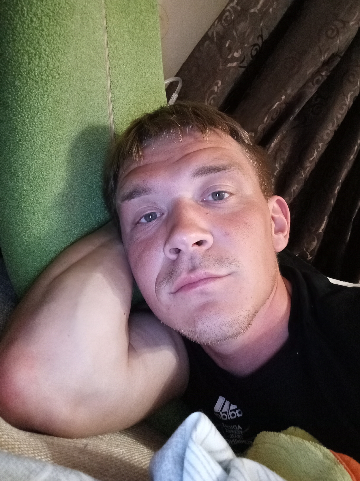 A male individual is depicted in a close-up, indoor image taken in Chaikovsky, Russia. The man, an adult with light skin, light brown hair, and a short goatee, is seen from the chest up, looking directly at the camera with a neutral expression. His head is supported by or resting against a bright green, textured fabric object, consistent with a pillow or cushion. His left arm is bent, with the forearm and elbow visible, positioned between his head and the green fabric. He wears a black t-shirt featuring a white Adidas logo on the chest. The background on the right side reveals dark brown curtains with a swirling, lighter pattern. In the lower foreground, a crumpled light-colored fabric object, possibly a towel or blanket, is visible. The scene is adequately lit, with a noticeable reflection on the man's forehead and nose.