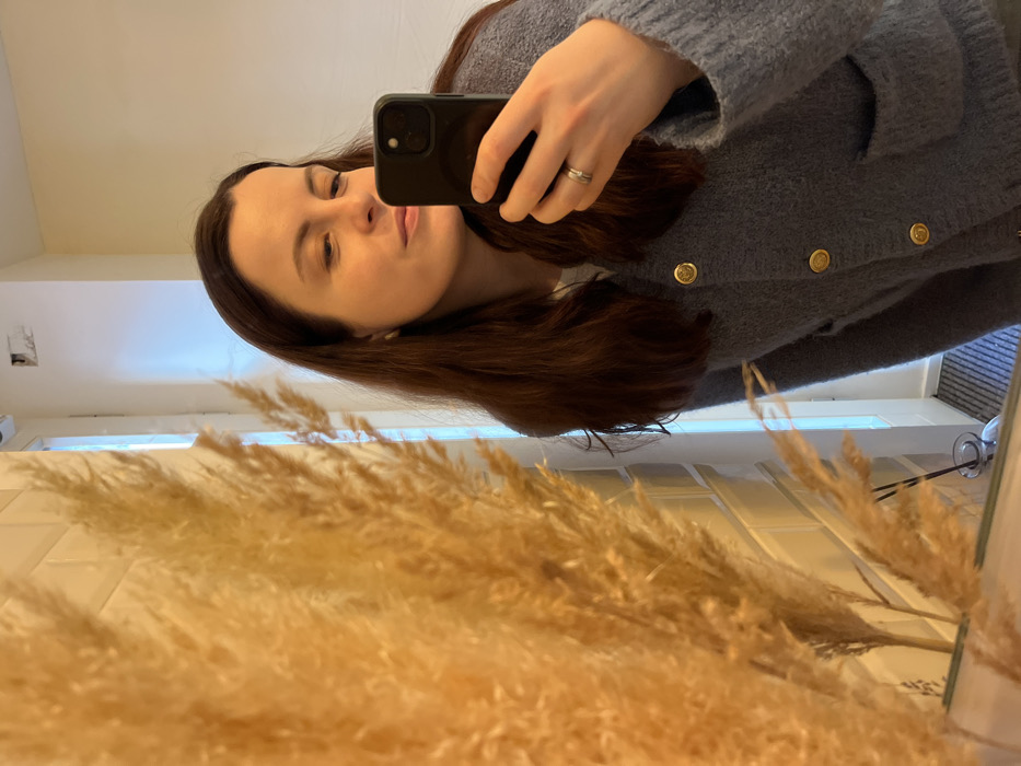 An adult female, reflected in a mirror, captures a self-portrait using a black smartphone held in her right hand. She has long, dark brown hair, fair complexion, and wears a light blue/grey textured knit cardigan with gold-colored circular buttons and two front pockets over a white garment. A two-band silver-colored ring adorns her left ring finger. The left foreground of the reflection is dominated by an out-of-focus arrangement of dried, yellowish-brown pampas grass. The background reveals a bright, minimalist interior with light-colored walls, possibly white rectangular tiles. A tall, narrow window or glass-paneled door frame is visible, admitting light with a blue hue, and a dark, striped floor mat lies near a threshold. A small glass diffuser bottle is reflected on a surface beneath the pampas grass. A ceiling-mounted device, resembling a security camera, and a minor wall imperfection are visible in the upper reflected background. This interior scene is located within Kaiserslautern, Germany, consistent with a modern commercial space such as a cafe or boutique.