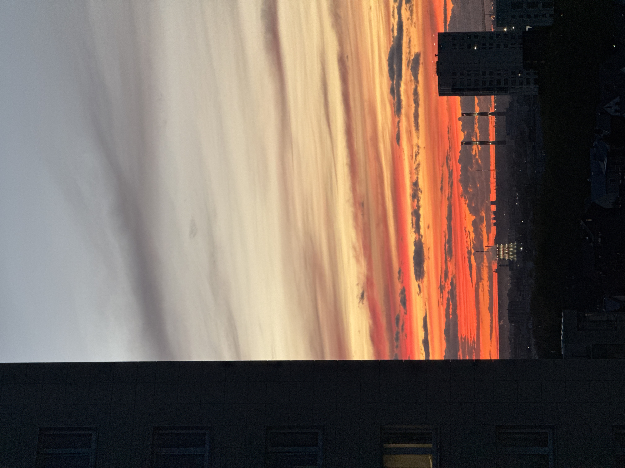 The image shows a cityscape at dusk in Kazan, Russia. The sky is filled with streaks of orange, red, and yellow light from the setting sun, contrasting with patches of grey and dark clouds. A tall building with multiple windows is visible on the right side of the frame. In the foreground, the top of another building, dark and silhouetted, is visible with a few lit windows. The location can be inferred from the architecture and urban setting consistent with Kazan.