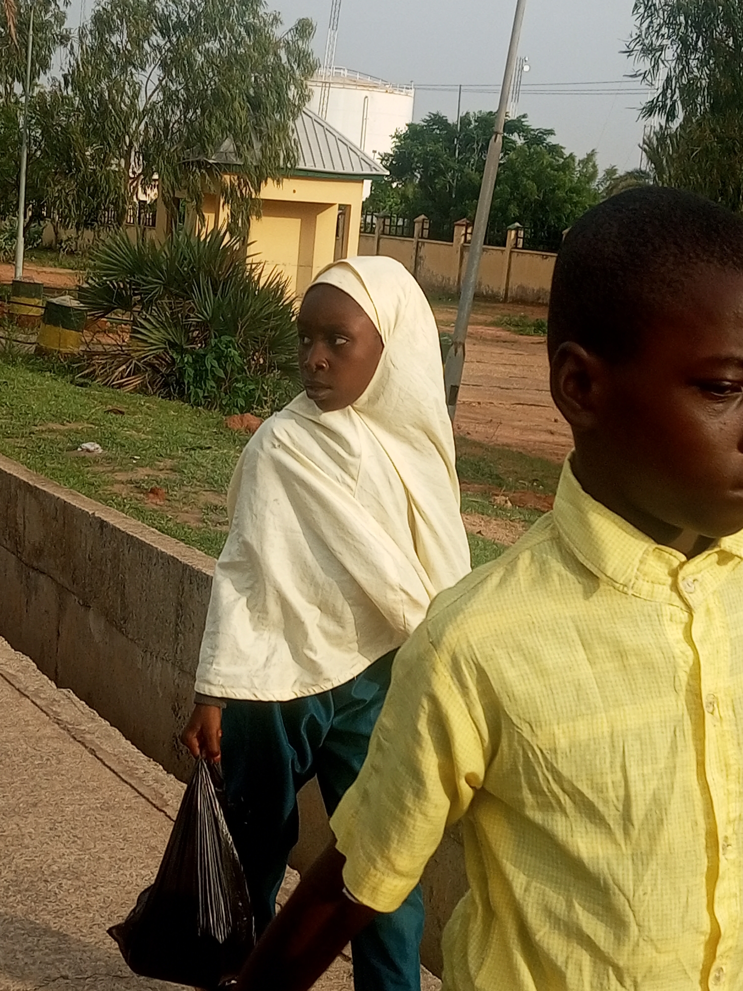This is a medium-shot, outdoor photograph taken during the daytime in Gombe, Nigeria. The image features two young individuals, a girl and a boy, positioned in the foreground. The girl, to the left of the frame, is wearing a light-colored headscarf and a dark teal garment, and she is carrying a black plastic bag in her left hand. She is looking to her left, away from the camera. The boy, to the right of the frame, is partially visible and is wearing a yellow checkered short-sleeved shirt. He is looking to his right, also away from the camera.
The background suggests an urban or semi-urban environment, possibly near a public facility or institution. Behind the children, there is a low concrete wall, with grassy and dirt areas beyond it. Further in the background, a light yellow building with a gray roof is visible, along with trees and fencing. A tall white structure, possibly a water tower or storage tank, can be seen in the distance. The overall lighting indicates it is likely late afternoon or early morning, given the warm tones and the shadows. The scene appears to be a moment captured in transit or while walking, with the children seemingly engaged in their own directions.