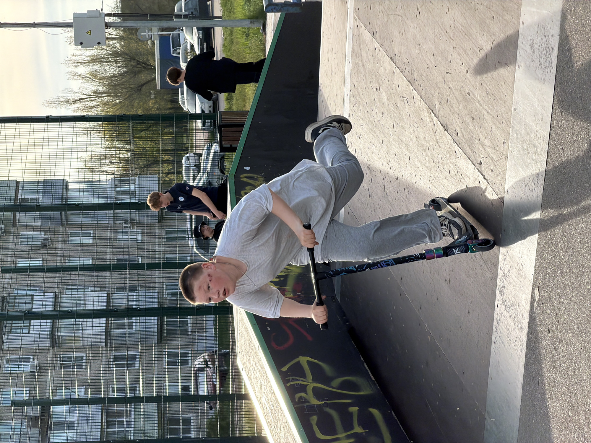A young person is riding a scooter on a skate ramp within a skate park. The skate park is enclosed by a tall, green-coated chain-link fence. Behind the fence is a multi-story residential building and several parked cars, possibly in a parking lot. The rider wears a light-colored t-shirt, gray sweatpants, and sneakers. There are other people visible in the background, one wearing a black jacket and another wearing a dark shirt and kneeling. The location is Kamyshin, Russia.