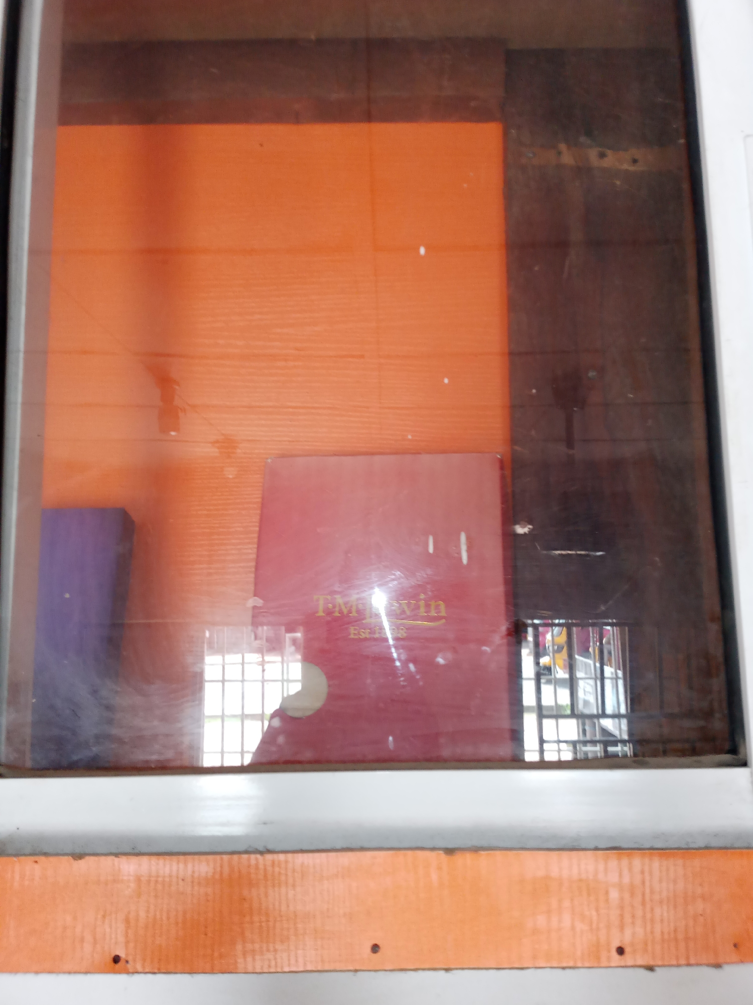 The image shows a close-up view of a glass display case. The background is a bright orange wall with a textured finish. A dark wooden panel is visible on the right side of the display case, showing some wear and tear.
Inside the case, a red rectangular object is the most prominent item. It appears to be a book or a box with text printed on it. The visible text reads "TM WIN" in a golden color, and below that, "Est. 1998". To the left of the red object, a purple rectangular object is partially visible. Reflected in the glass, a grid-like pattern is seen, likely from a window or a light fixture. There are also smudges and dust marks on the glass surface.
The overall scene suggests an indoor display, possibly in a shop or a public space. The lighting appears to be natural, given the reflections, and the time of day is indeterminate. The location context of Jalingo, Nigeria, doesn't provide specific visual cues within the image itself, but places the scene within that geographical area. No people are visible in the image.