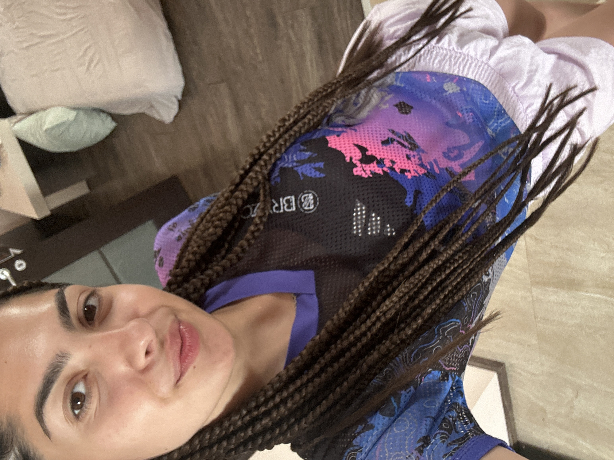 A young woman with long, dark braided hair is taking a selfie. She is wearing a purple and blue athletic top with a graphic print and light purple athletic shorts. Her face is looking towards the camera with a slight smile. The background appears to be an indoor setting with wooden flooring, a bed with a light pink comforter, and a white nightstand. The scene is lit by artificial light.