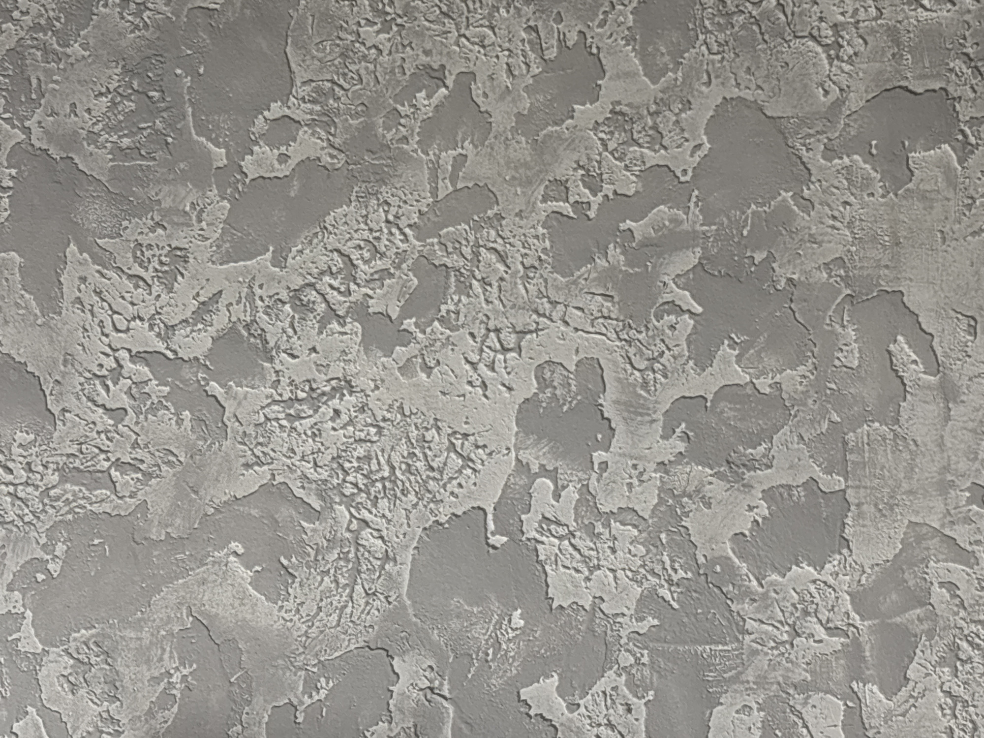 The image displays a vertically oriented, close-up view of a textured surface, likely a decorative wall finish, located in Unknown, Unknown. The surface exhibits a pronounced, irregular relief pattern, characterized by areas of rough, lighter grey material elevated above smoother, darker grey depressions. The pattern is organic and abstract, resembling a mottled or stucco-like application with varying degrees of coarseness and depth. The illumination creates subtle shadowing that accentuates the three-dimensional quality of the texture. No people, objects, actions, or further environmental details are present.