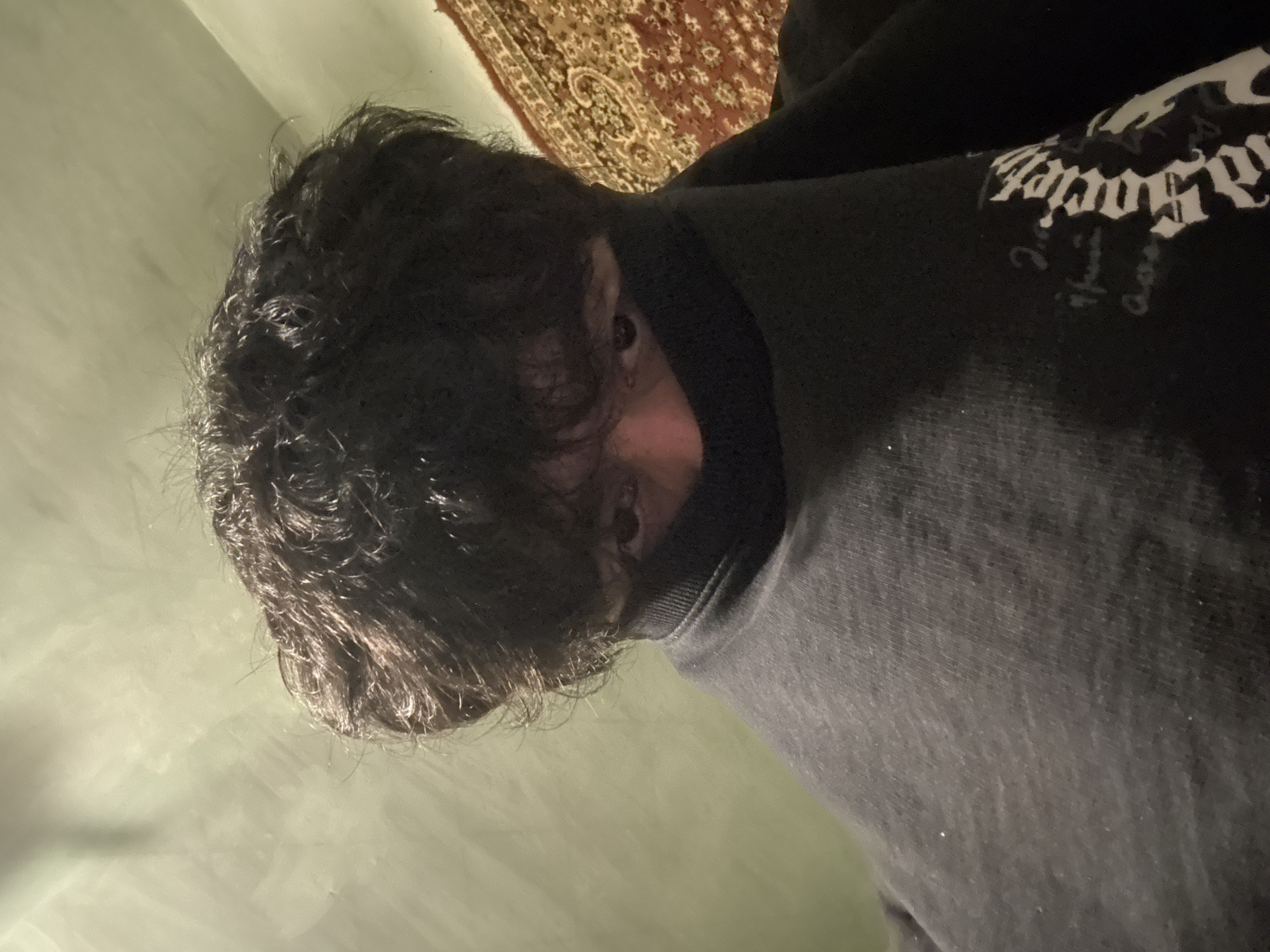 A photograph shows a person with dark, wavy hair wearing a black turtleneck sweater or sweatshirt with white lettering on the upper right. The face is partially obscured by hair and the high collar of the garment, but the eyes are visible. Behind the person is a carpet with an intricate pattern of brown, tan, and red. The background is a pale green wall. There is no information to determine the location in the city of Yerevan, Armenia.