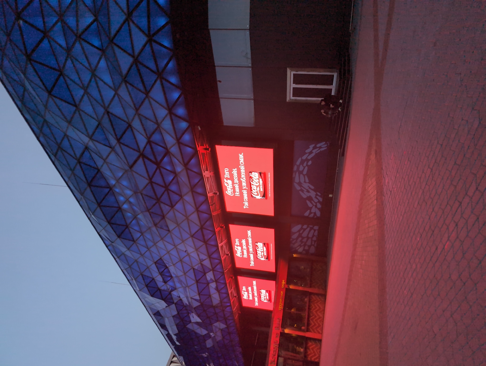 The image depicts a modern building with a distinctive faceted blue glass facade under a dusky sky. Below the glass facade, a row of three large, bright red digital billboards displays advertisements for Coca-Cola Zero, featuring the brand's logo and slogans in Ukrainian. The billboards are illuminated, casting a red glow onto the ground and a building entrance below. To the left of the billboards, a more traditional building facade with signs is visible, including one that appears to read "Georgian Cafe". In the foreground, a paved area, possibly a plaza or walkway, is lit with a reddish hue from the billboards and ambient lighting. Two individuals are seated on a set of steps leading to a white-framed door on the right side of the image. The overall scene suggests an urban environment at dusk, likely a commercial or entertainment area. Based on the presence of Ukrainian language on the advertisements and the architectural style, the location is identified as Kyiv, Ukraine.