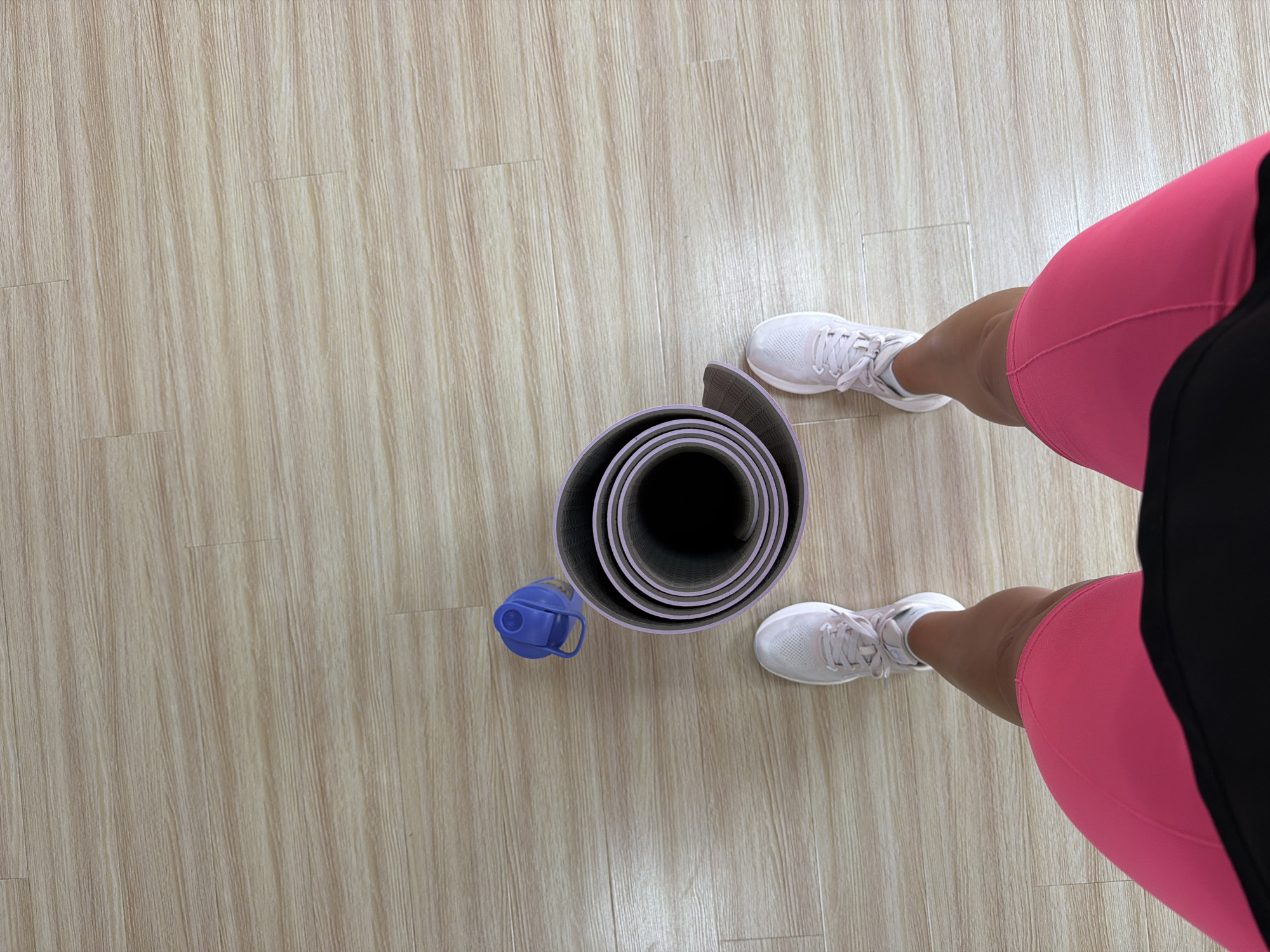 An overhead, full-body shot of a person standing on a light wood-grain floor, ready to exercise. The person is wearing pink athletic shorts, white sneakers, and a black top. To their left, on the floor, is a rolled-up purple yoga mat and a blue water bottle. The scene takes place indoors, likely in a home or gym setting. There is no indication that this scene is in Sochi, Russia.