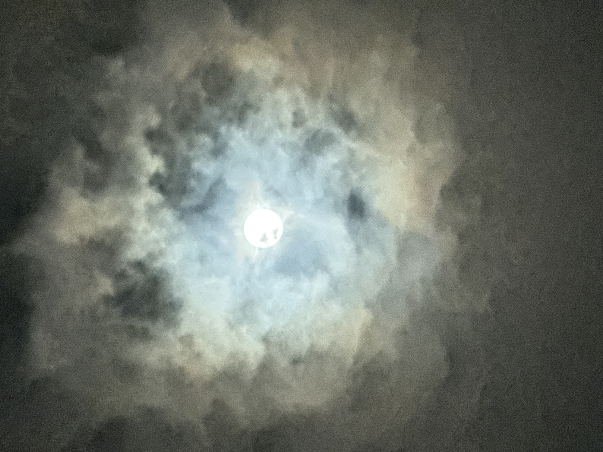 The image depicts the moon partially obscured by clouds at night. The moon is a bright, luminous orb positioned slightly off-center towards the top left of the frame. It is surrounded by wispy and dense clouds, which create a halo effect and obscure parts of the lunar surface. The clouds vary in color from bright white, where they are illuminated by the moon, to dark grays and blacks, indicating their density and the absence of direct moonlight. The overall impression is one of a cloudy night sky, with the moon struggling to break through the atmospheric cover. The lighting suggests it is nighttime, and the presence of clouds indicates overcast or partly cloudy weather. There are no visible people, objects, text, or specific location cues within the image itself, other than the context provided that it was taken in Nha Trang, Vietnam. The mood conveyed is one of mystery and ethereal beauty, with the obscured moon lending a dramatic and somewhat somber atmosphere.