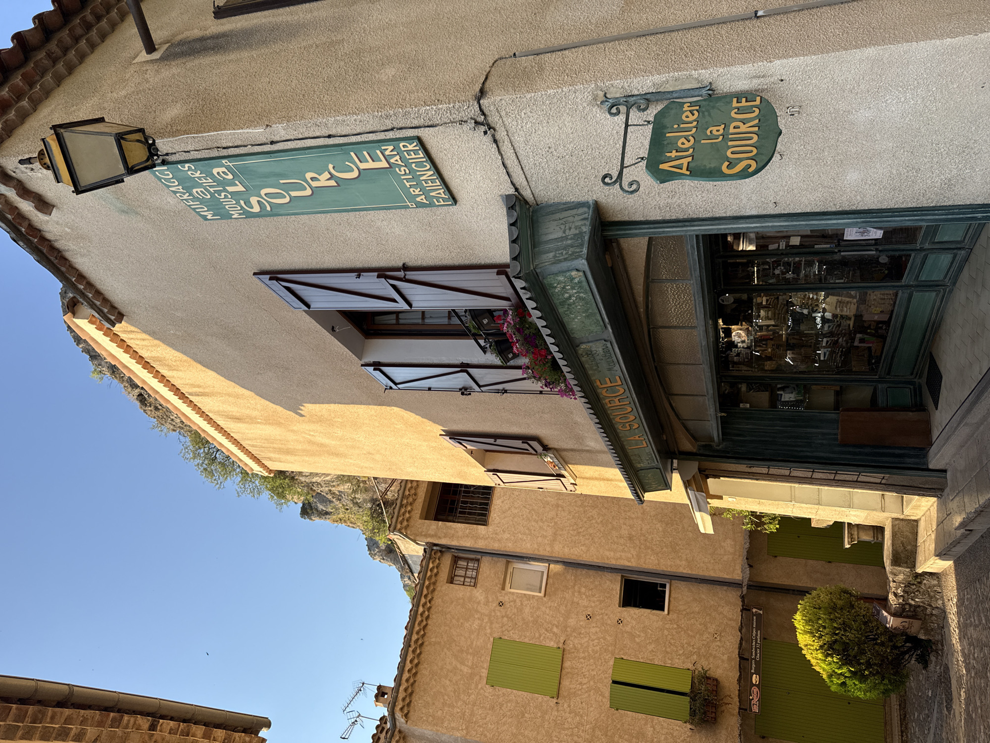 The image depicts a narrow cobblestone street in the village of Moustiers-Sainte-Marie, France, bathed in the warm light of late afternoon. The buildings are constructed of pale, textured plaster and feature terracotta tiled roofs. The focal point is a shop with a green-framed glass storefront, displaying various items inside. Above the shop entrance, a green sign reads "LA SOURCE." A vertical green sign on the building's corner, adorned with a streetlamp, displays "MUFRAGGI A MOUSTIERS La SOURCE ARTISAN FAIENCIER." Another hanging green sign reads "Atelier la SOURCE." Shutters on the buildings are a mix of dark wood and bright green. A small, leafy green bush grows from a stone planter on the left. The background reveals a rugged, rocky cliff face. The sky is a clear, vibrant blue.