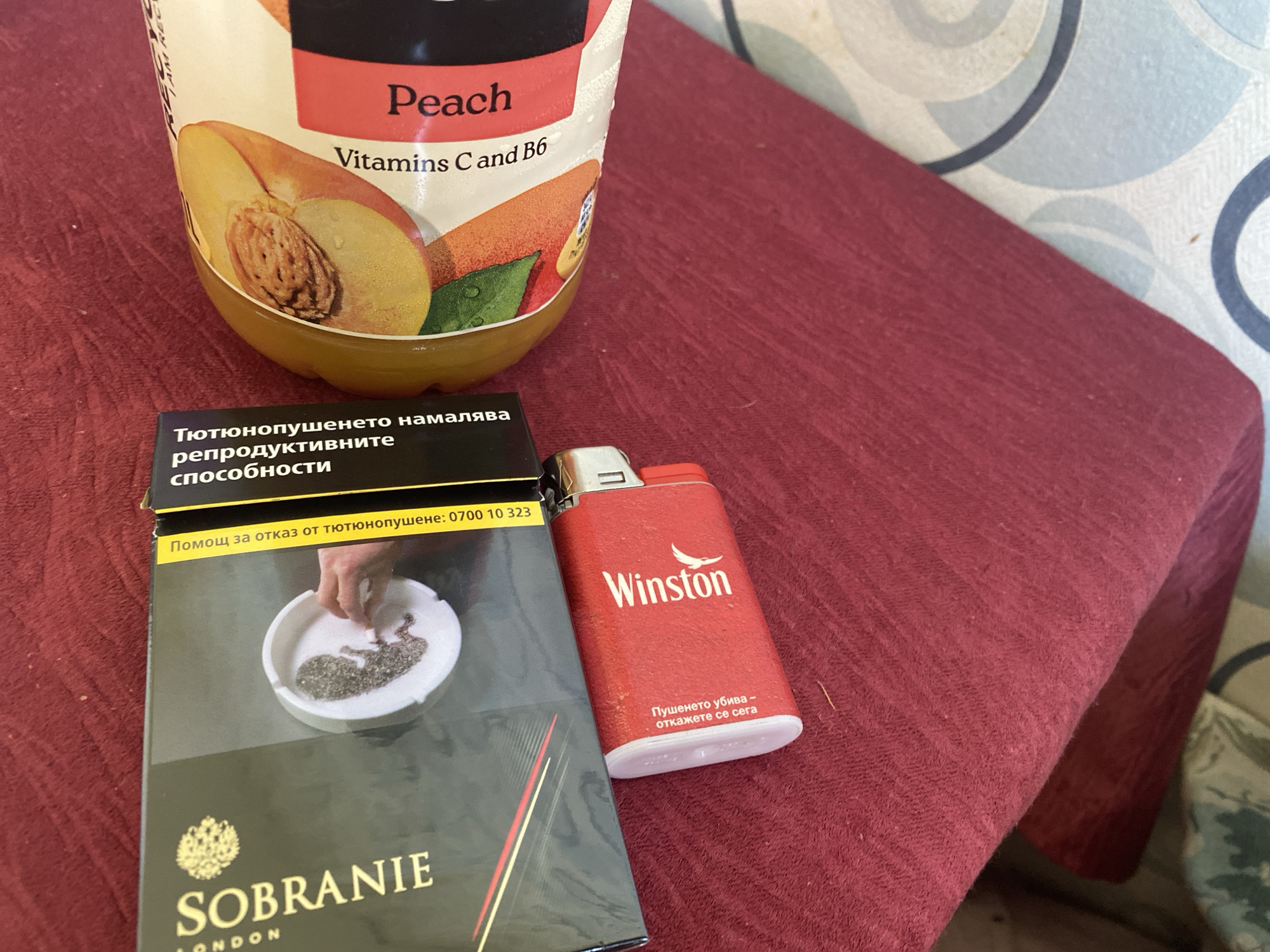 An eye-level shot captures a still life arrangement of a juice bottle, a cigarette pack, and a lighter resting on a textured, deep red fabric surface, possibly a tablecloth or blanket. In the background, a light blue and grey patterned surface is visible, suggesting a wall or another piece of furniture.
The prominent object on the left is a bottle of peach-flavored juice, labeled "Peach" with "Vitamins C and B6" indicated. An image of a halved peach with its pit is visible on the label. To the right and slightly below the juice bottle is a pack of "SOBRANIE LONDON" cigarettes. The pack features a graphic health warning depicting a hand shaping what appears to be a fetus out of ash in an ashtray. Text on the pack, in Bulgarian, reads: "Тютюнопушенето намалява репродуктивните способности" (Smoking reduces reproductive ability) and "Помощ за отказ от тютюнопушене: 0700 10 323" (Help to quit smoking: 0700 10 323). Next to the cigarette pack is a red lighter bearing the "Winston" brand name. It also displays a Bulgarian warning: "Пушенето убива - откажете се сега" (Smoking kills - quit now).
The lighting is soft and even, typical of an indoor setting, and does not suggest a particular time of day. No specific activity is depicted, but the items suggest a moment of relaxation or a break.