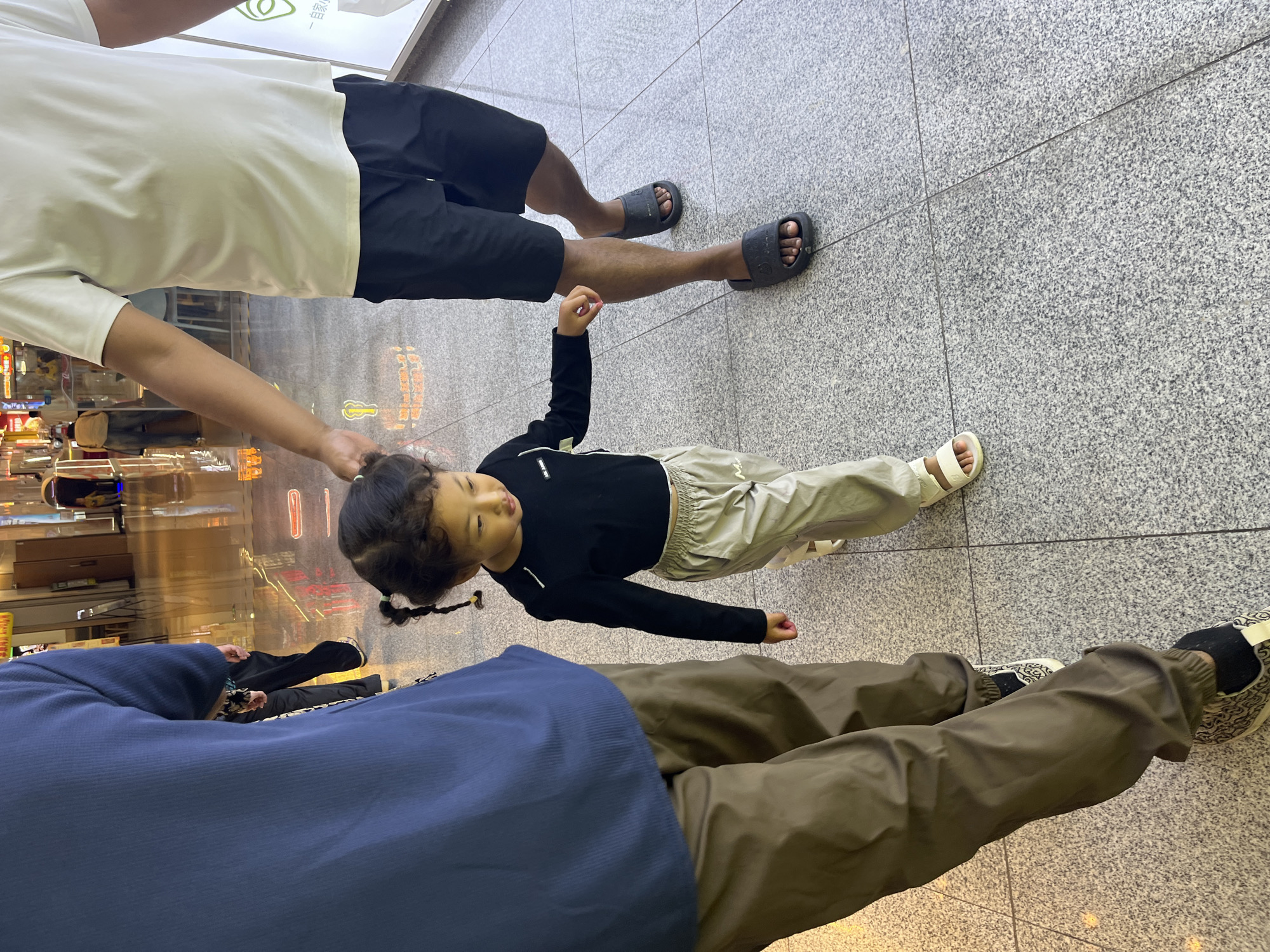 The image depicts three people standing on a tiled floor, likely indoors in a commercial setting such as a shopping mall or airport, given the reflections of lights and signage in the background. The perspective is from a low angle, looking up at the subjects.
In the foreground, a person is wearing a navy blue top and brown pants, with one leg bent. To their left and slightly behind, another person is wearing a white t-shirt and black shorts, with their legs extended. Between these two individuals, a young child stands facing towards the camera. The child is wearing a black long-sleeved shirt and light-colored pants. The child has dark hair styled in pigtails and appears to be looking upwards and slightly to the side with a neutral expression.
The floor is a light gray speckled tile with visible grout lines. The background is blurred, showing reflections that suggest a bright, possibly evening or interior lighting environment. There are no obvious signs of weather conditions, but the indoor setting implies controlled climate. The overall scene captures a moment of pause or waiting, with the people standing relatively still. No discernible text is visible on the clothing or in the immediate surroundings of the people, although faint illuminated signs are present in the distant background.
