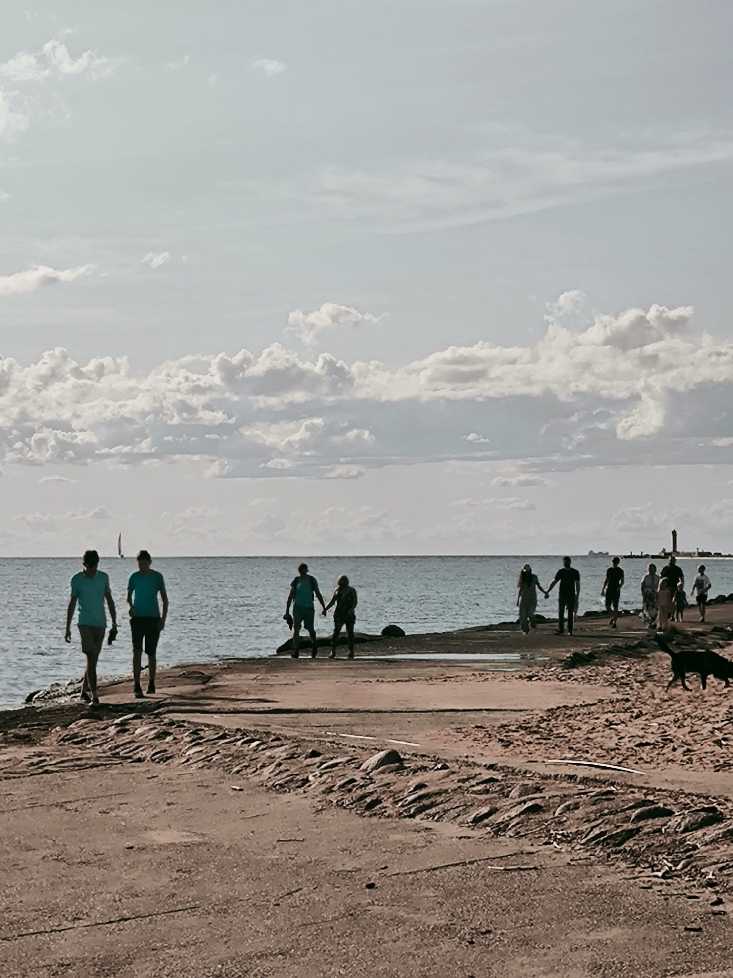 A group of people are walking on a pier by the sea in Riga, Latvia. Two men in blue t-shirts are walking towards the viewer, while a group of people are walking away from the viewer further down the pier. A black dog is running on the sand in the foreground. The sky is cloudy and the sea is calm. A lighthouse and a sailboat are visible in the distance. The pier appears to be made of concrete and is lined with rocks.
