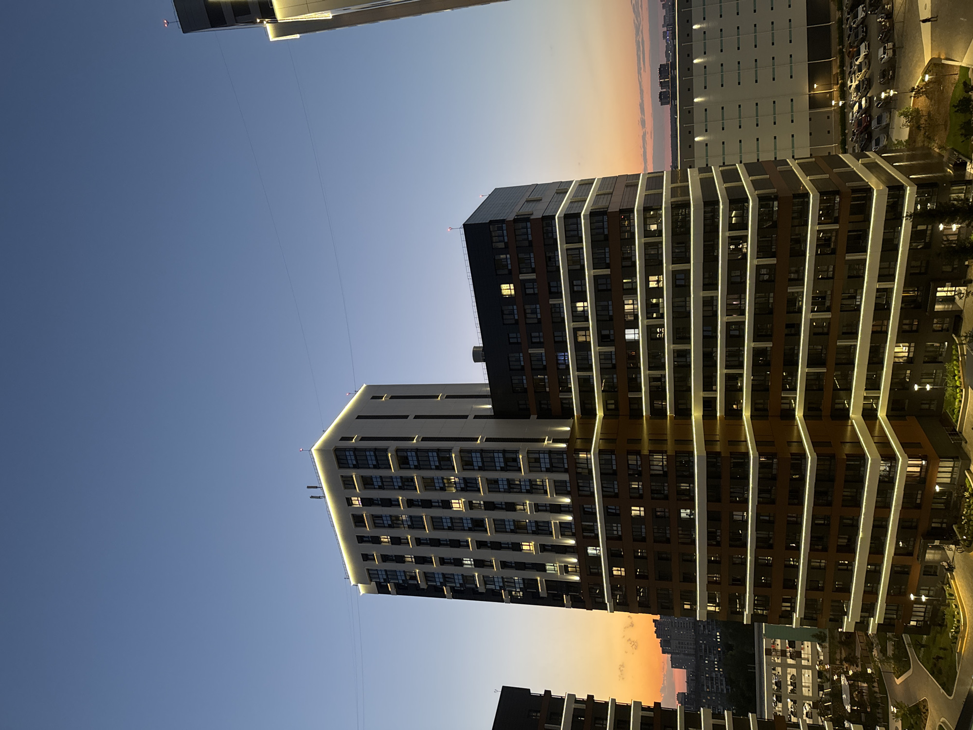 An exterior, high-angle shot captures a modern apartment complex at dusk in Novosibirsk, Russia. The sky transitions from a deep blue at the top to a gradient of orange and pink near the horizon, indicating the setting sun. Two prominent, multi-story buildings dominate the foreground. The building on the left is a tall, rectangular structure with a facade of dark brown and cream-colored panels. It is illuminated by a series of warm, white lights outlining its horizontal edges and vertical corner accents, as well as lights in individual windows. Adjacent to it, to the right, is a slightly shorter, similarly designed building, also featuring brown and dark gray panels and the same illuminating lighting scheme.
In the middle ground, a large, flat-roofed building with a white facade and a grid of lights is visible, likely a commercial or industrial structure. A parking lot filled with numerous cars is situated in front of this building. The background shows a city skyline with more buildings, some of which are illuminated. The overall scene depicts a contemporary urban environment at twilight, with the architecture of the apartment complex highlighted by artificial lighting against the natural fading light of the day.