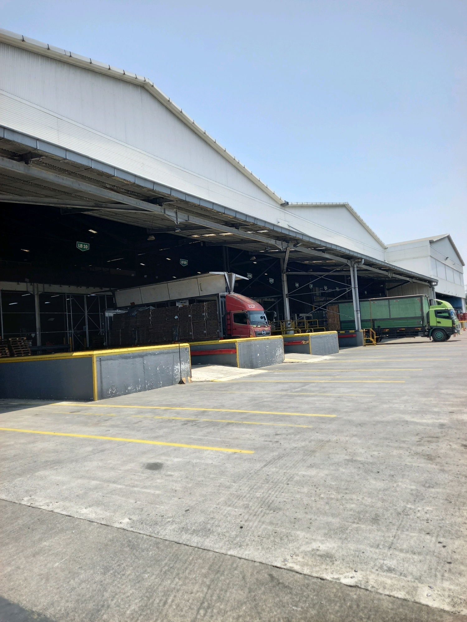 The image depicts an outdoor loading dock area at a warehouse facility in Gempol, Indonesia. The scene is dominated by a large, corrugated metal industrial building with a wide, covered overhang. The ground is paved with concrete, marked with yellow parking lines.
In the foreground, a raised loading dock platform is visible, painted with yellow and gray markings. Pallets are stacked on the left side of the platform. Two trucks are positioned at the loading dock. On the left, a red truck with the branding "DAYA" is parked, partially obscured by the loading dock. On the right, a green truck is backed up to the dock, its trailer open, suggesting loading or unloading activities.
Inside the covered area of the warehouse, several bays are marked with green signs, including "LB 10" and "LB 09". Metal beams and structural supports are visible within the warehouse. The sky above is clear and blue, indicating a bright, sunny day. The overall impression is a busy logistics or distribution center.